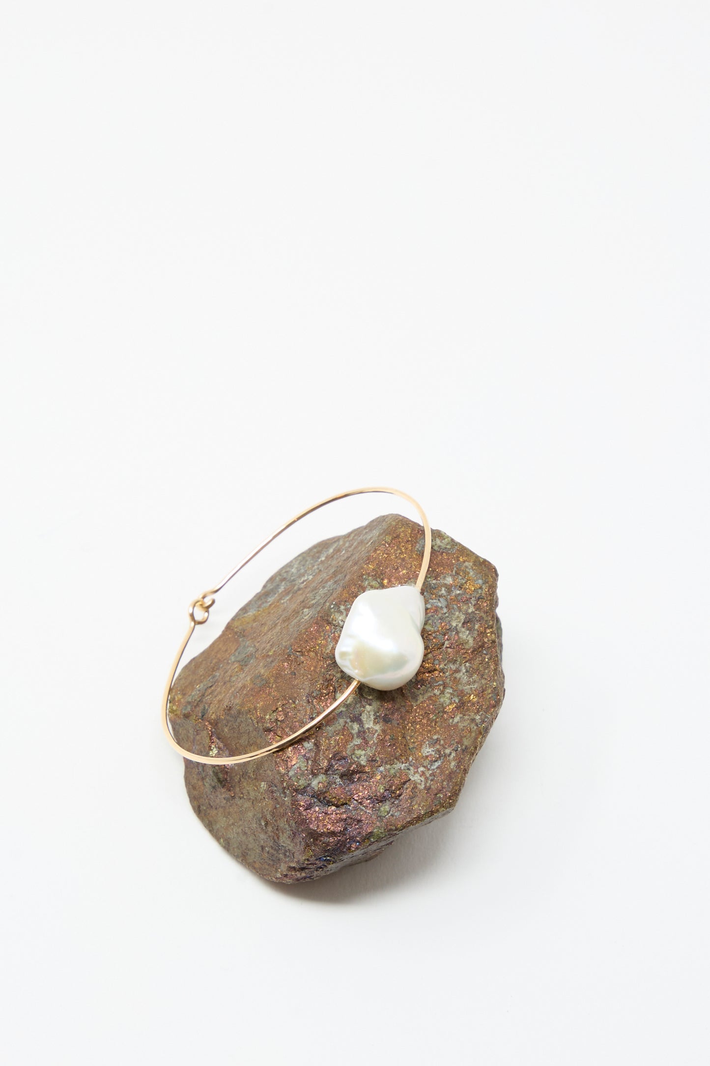 The Baroque Pearl Cuff Bracelet in White by Mary MacGill, featuring a single irregular white pearl on a gold-filled band, is elegantly showcased atop a rough brown stone against a plain white background.