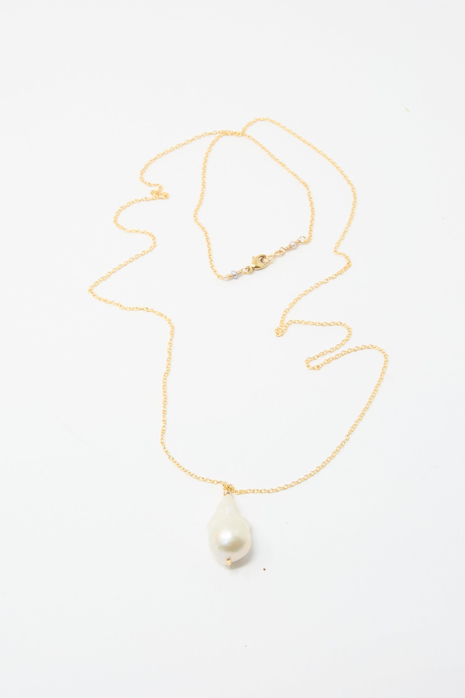 The Baroque Pearl Drop Necklace in White from Mary MacGill is beautifully showcased against a white backdrop, highlighting its handmade design with a gold chain and a single large Baroque pearl pendant.