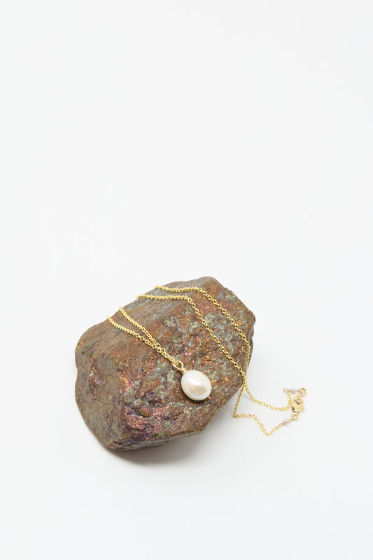 The Baroque Pearl Drop Necklace in White - 18" by Mary MacGill, featuring a single baroque pearl pendant on a gold chain, is displayed over an iridescent stone against a plain white background.