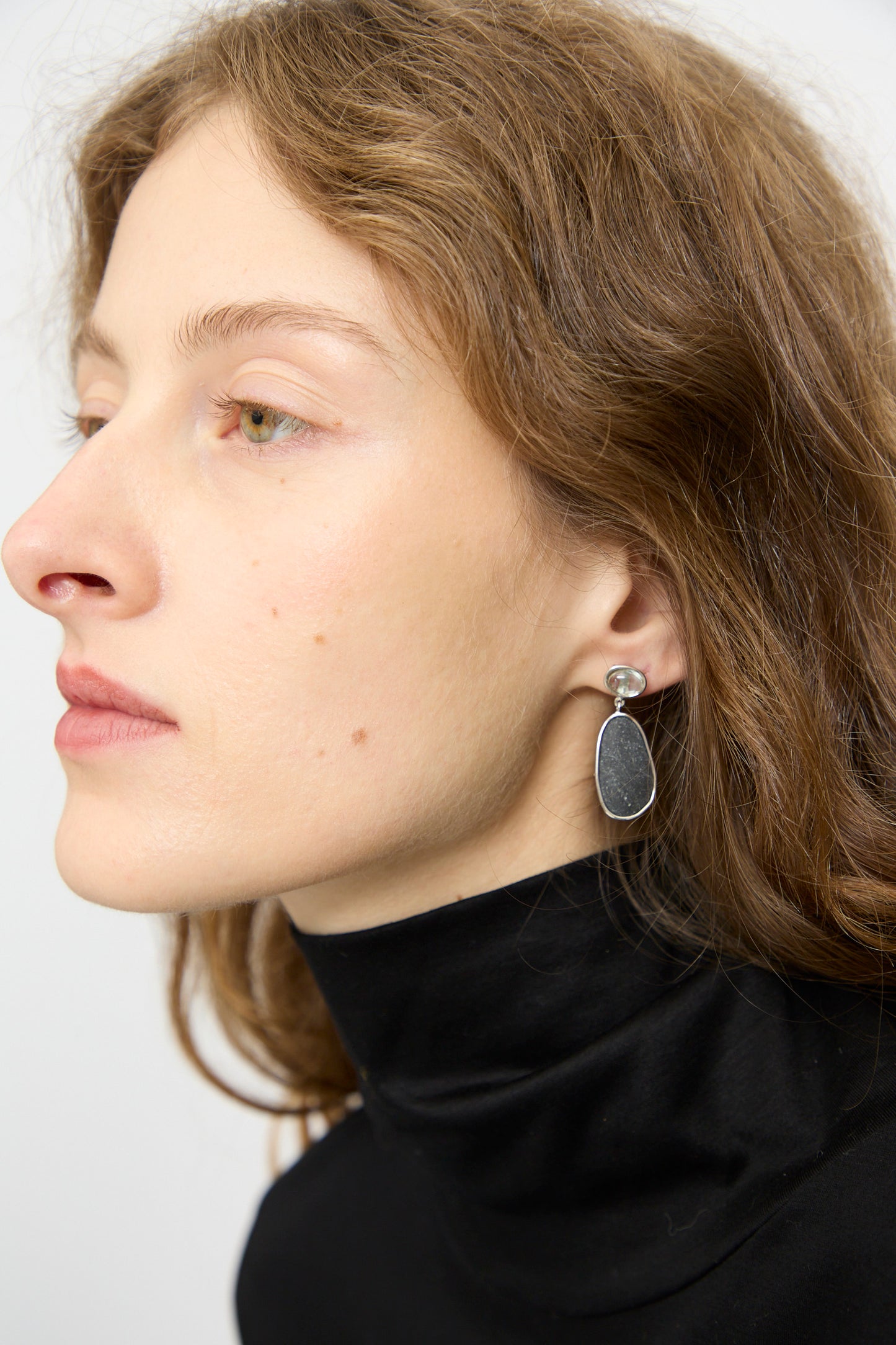 A woman with light skin and wavy brown hair wears a black turtleneck and Mary MacGill Double Stone Drop Earrings in Dark Island Stone and Tourmaline, featuring teardrop-shaped drops with round studs.