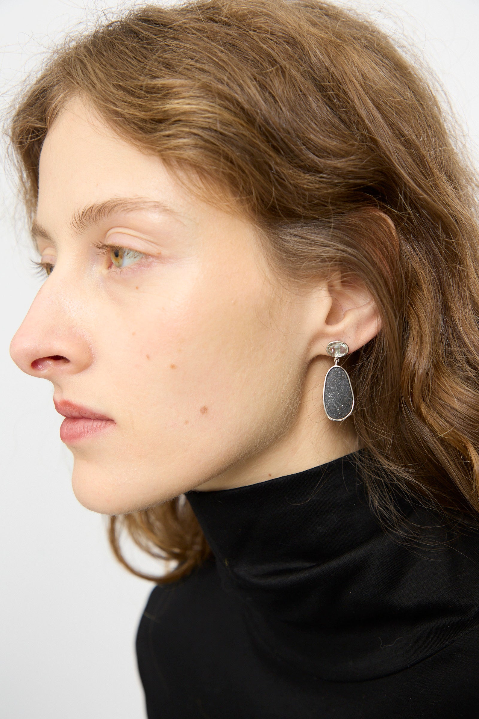 Woman with wavy brown hair in a black turtleneck wears Mary MacGill Double Stone Drop Earrings in Dark Island Stone and Tourmaline, shown in profile against a plain background.