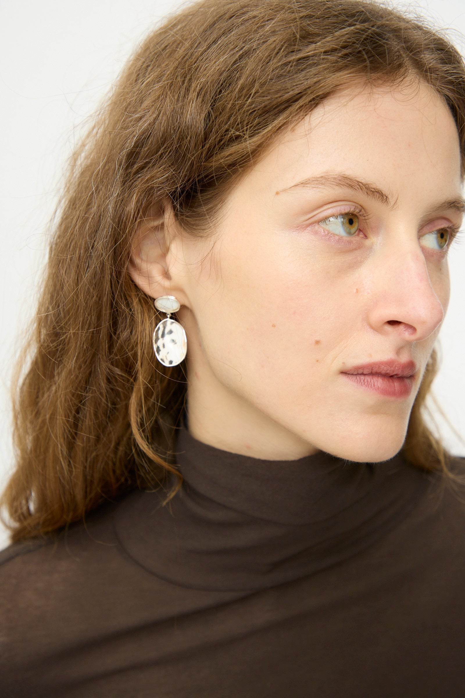 A woman with light brown hair in a brown turtleneck wears Mary MacGill Double Stone Drop Earrings in Moonstone and Cowrie Shell, gazing slightly to the side against a plain background.