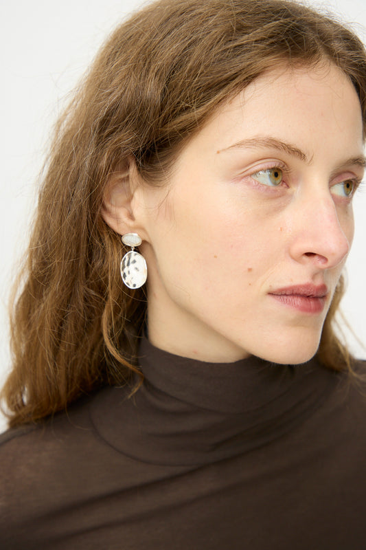 A woman with light brown hair in a brown turtleneck wears Mary MacGill Double Stone Drop Earrings in Moonstone and Cowrie Shell, gazing slightly to the side against a plain background.