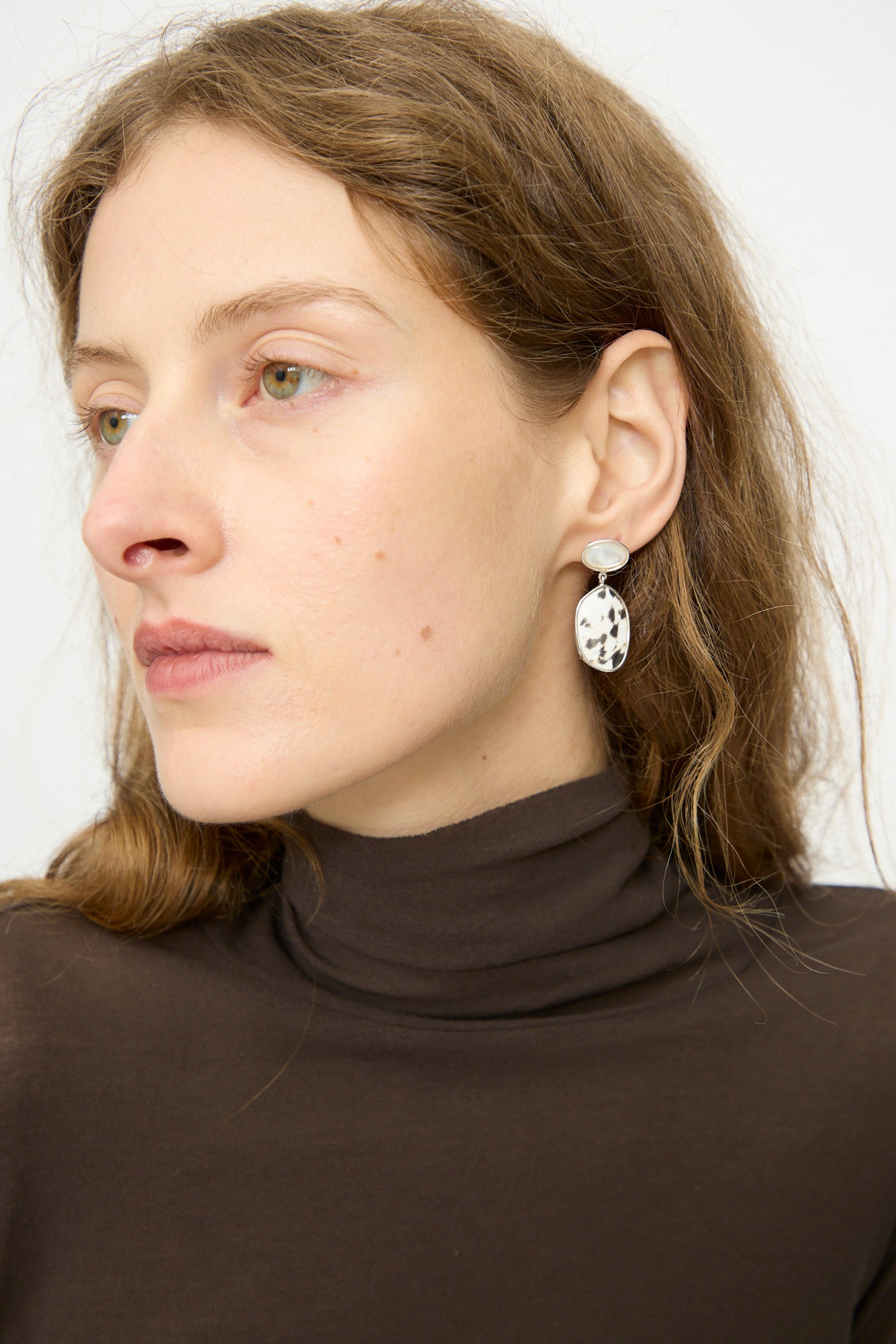 A woman with light brown hair wears a brown turtleneck and Mary MacGill Double Stone Drop Earrings in Moonstone and Cowrie Shell, featuring white stones with black speckles, looking to the side against a plain background.