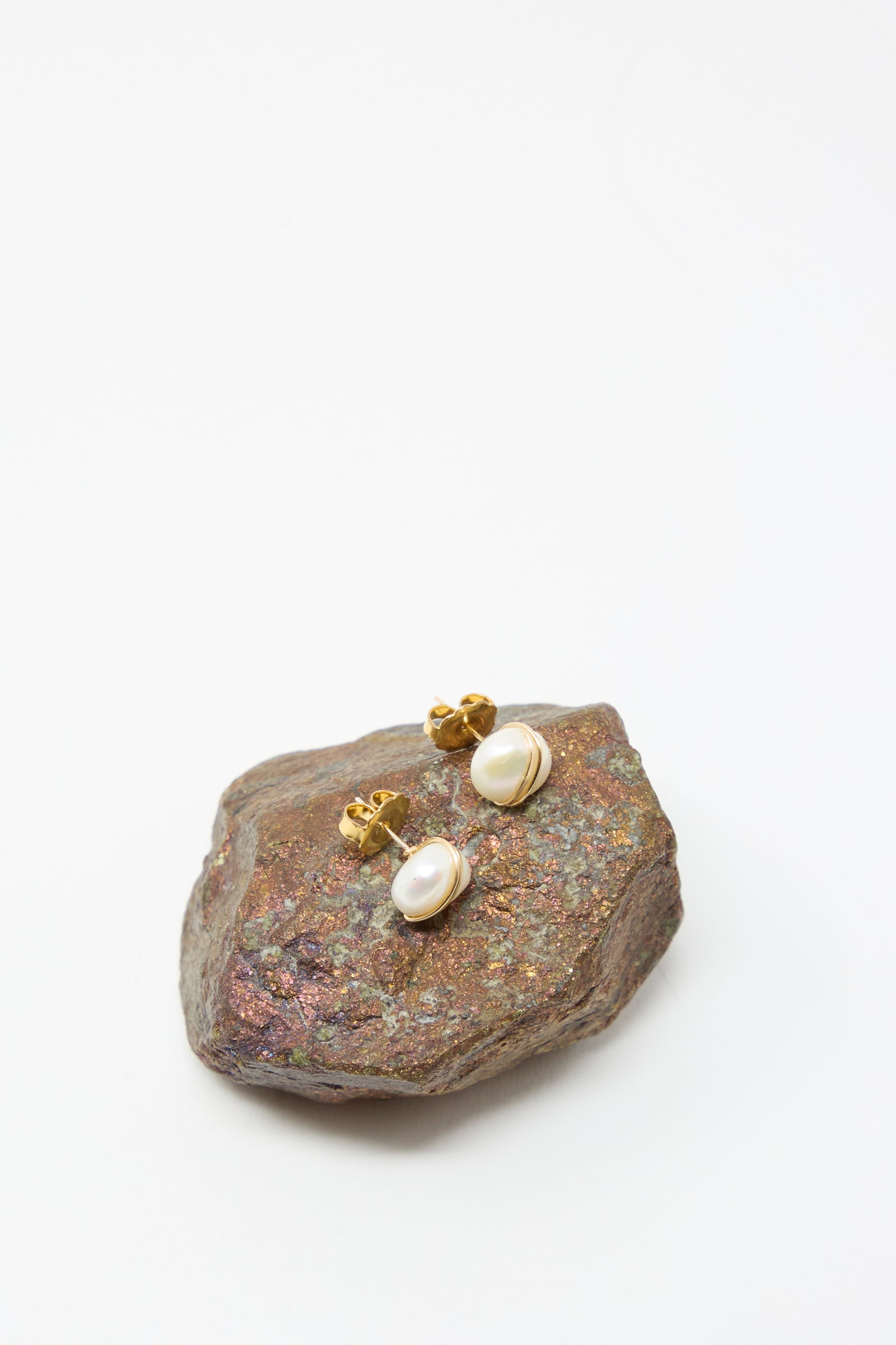 The Grande Baroque Pearl Studs in White by Mary MacGill feature handmade baroque pearls on gold-filled wire, shown on a textured brown rock with a white background.