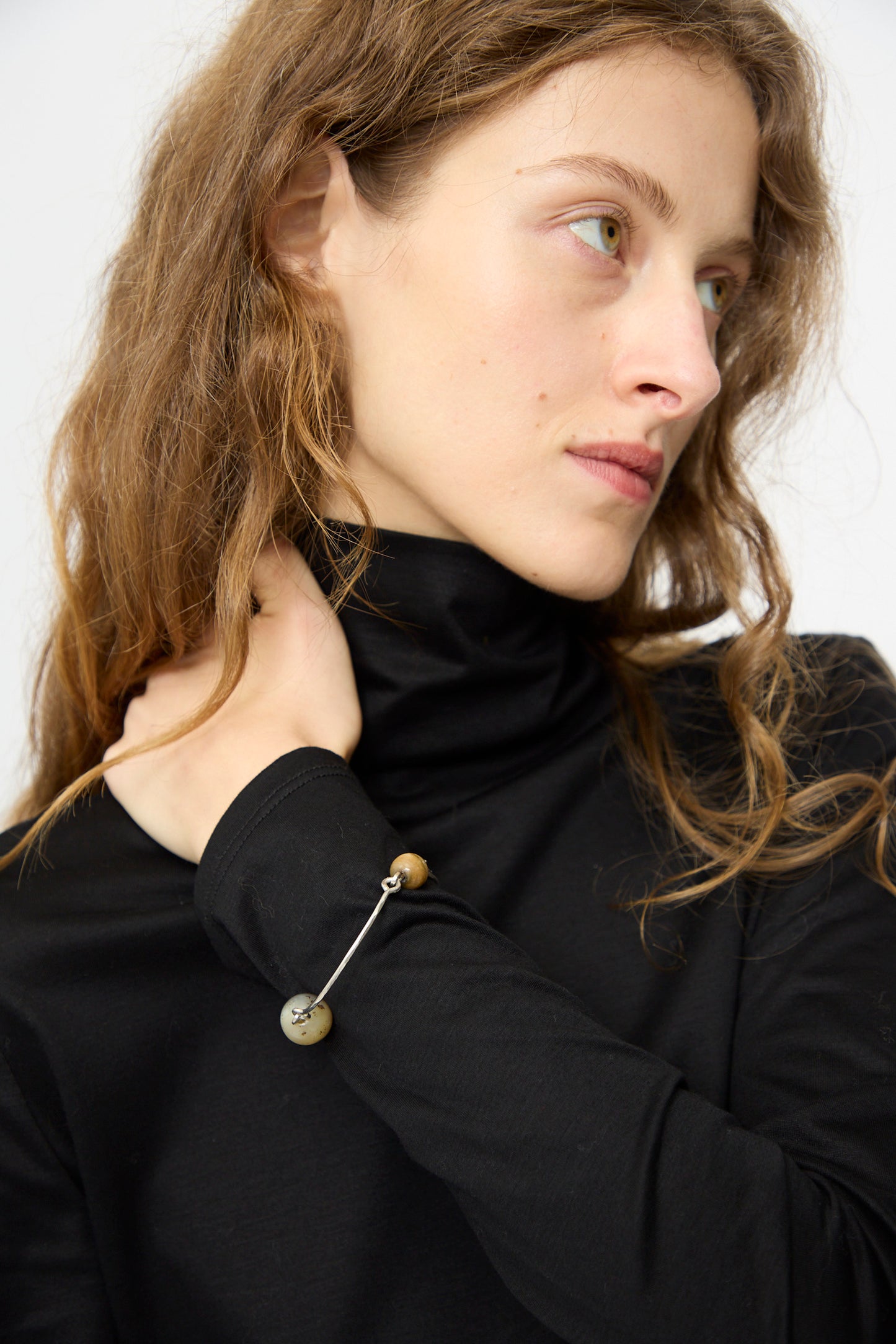 A woman with wavy light brown hair wears a black turtleneck and the Mary MacGill Hammered Sterling Silver Bangle with Ancient Agate Trade Beads, glancing to the side against a plain background.