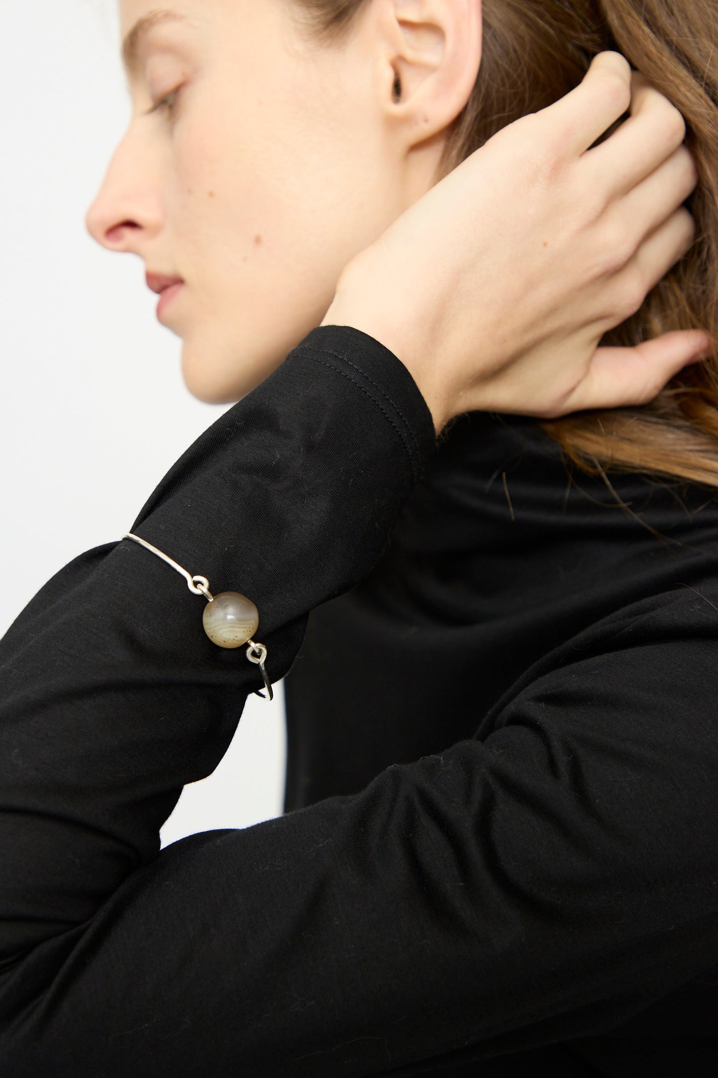 A person with light skin and brown hair wears a black long-sleeve top and the Mary MacGill Hammered Sterling Silver Bangle with Ancient Agate Trade Beads, highlighting their appreciation for unique handmade jewelry.