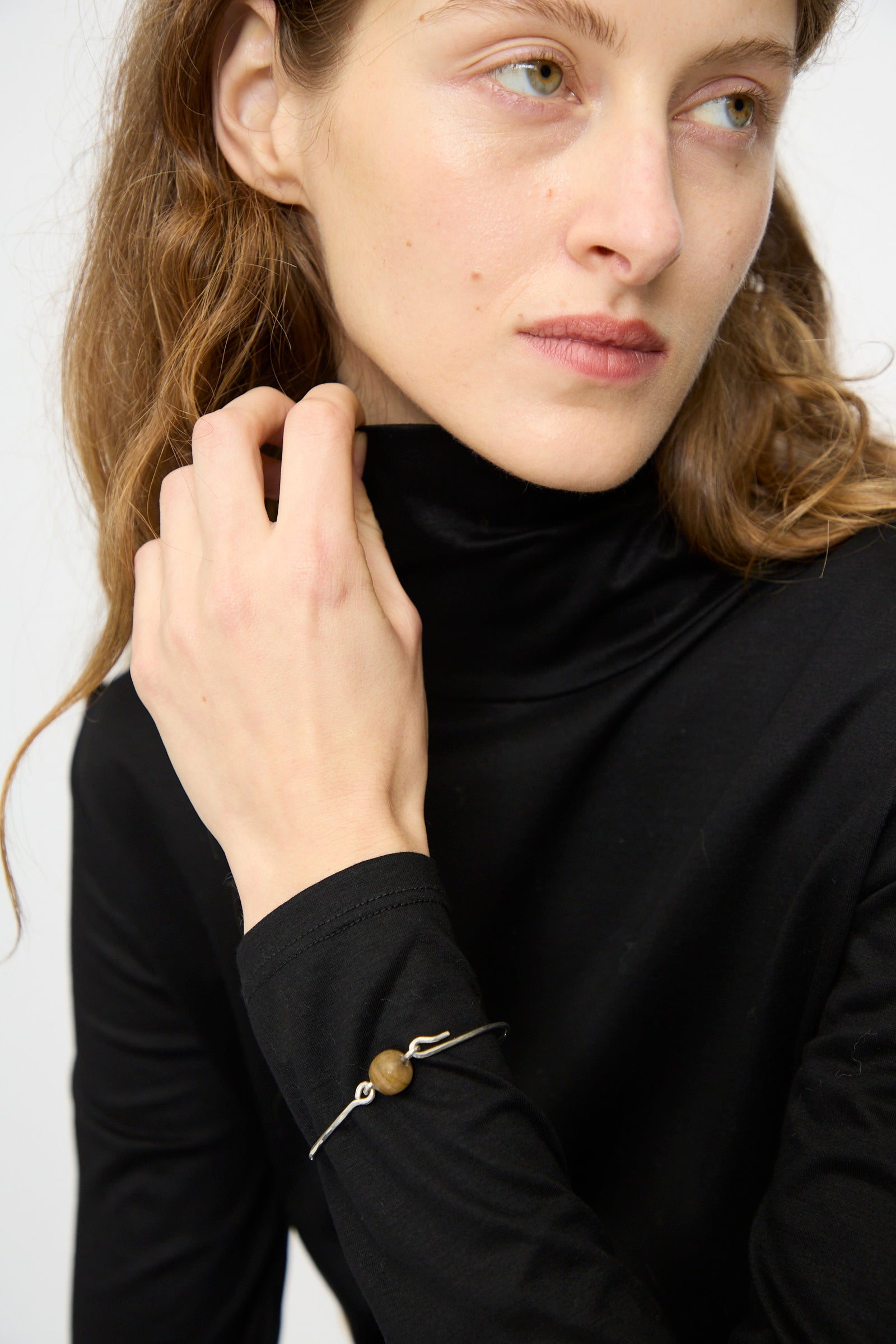 A person in a black turtleneck glances sideways, displaying the Mary MacGill Hammered Sterling Silver Bangle with Ancient Agate Trade Beads on their wrist.