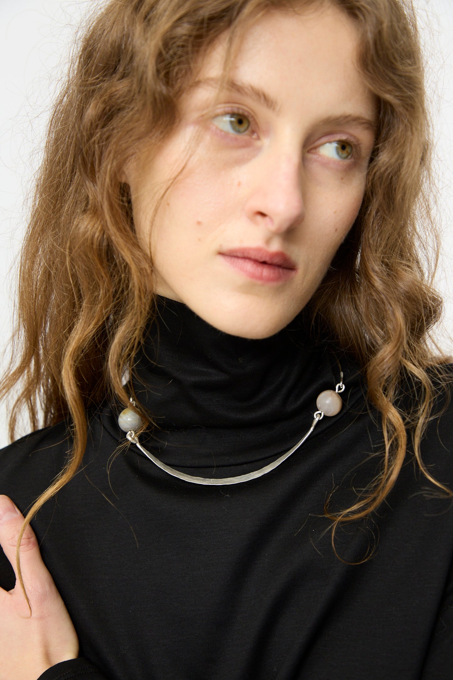 A woman with wavy light brown hair wears a black turtleneck and the Mary MacGill Hammered Sterling Silver Collar with Ancient Agate Trade Beads, gazing slightly to the side.