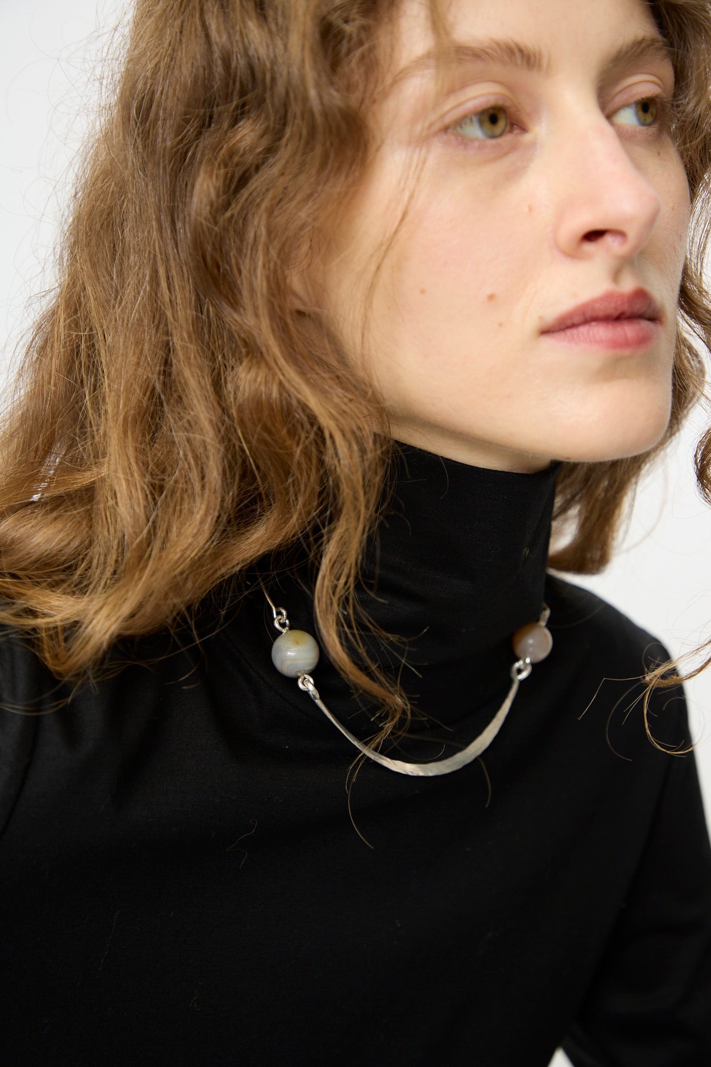 A person with long wavy hair in a black turtleneck wears the Mary MacGill Hammered Sterling Silver Collar with Ancient Agate Trade Beads, gazing slightly to the side.