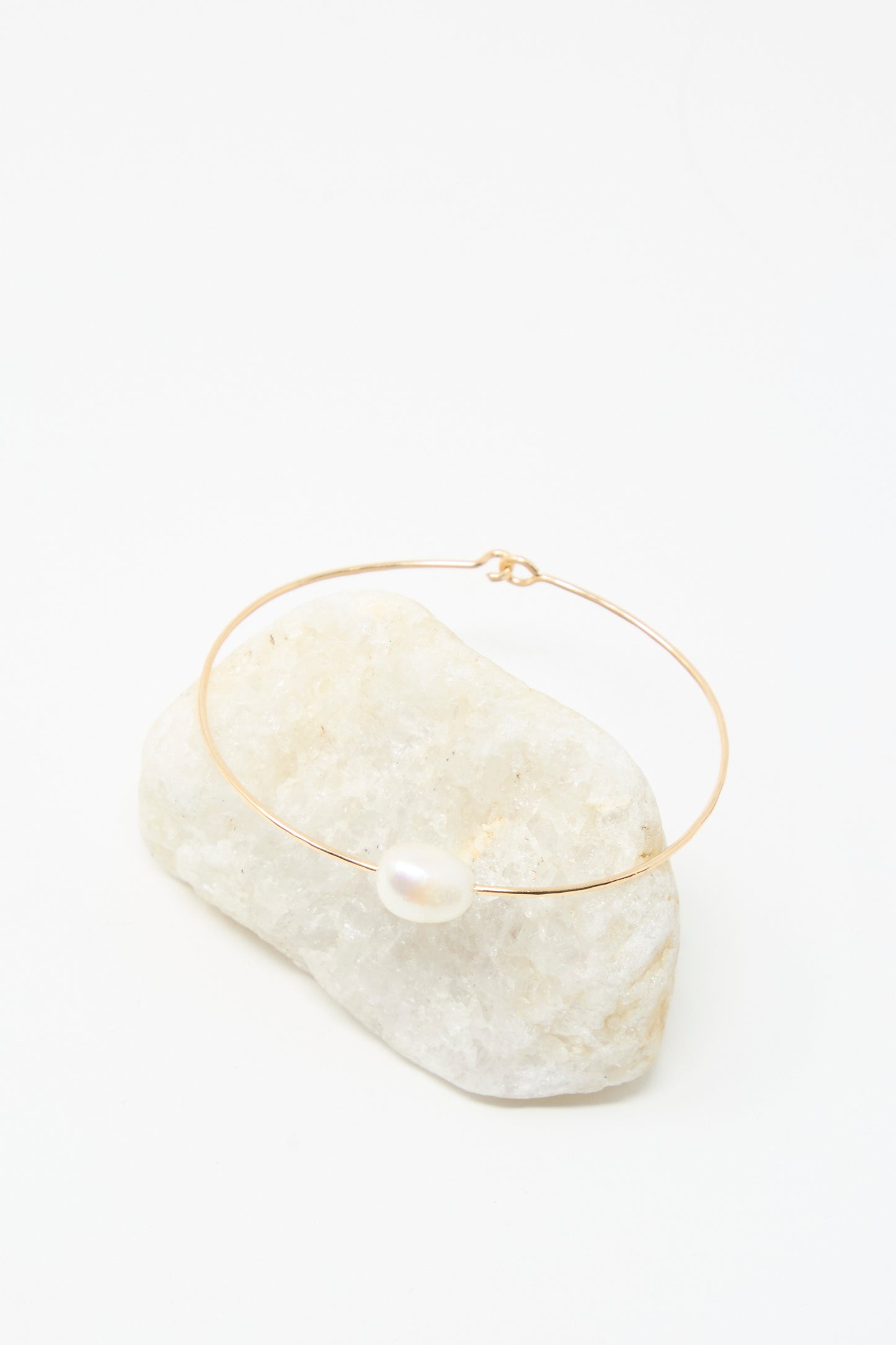 The Mary MacGill Petite Baroque Pearl Cuff in White, a delicate gold-filled bracelet featuring a single white pearl, is displayed on a white stone with a white background.