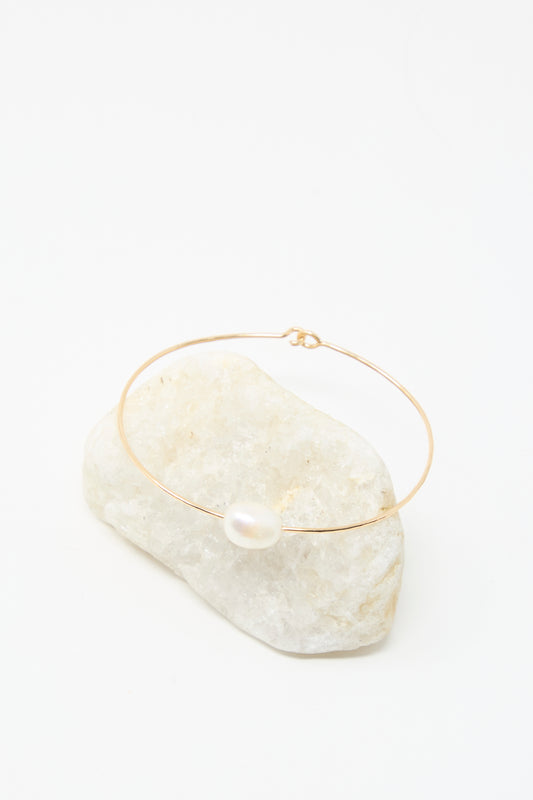 The Mary MacGill Petite Baroque Pearl Cuff in White, a delicate gold-filled bracelet featuring a single white pearl, is displayed on a white stone with a white background.