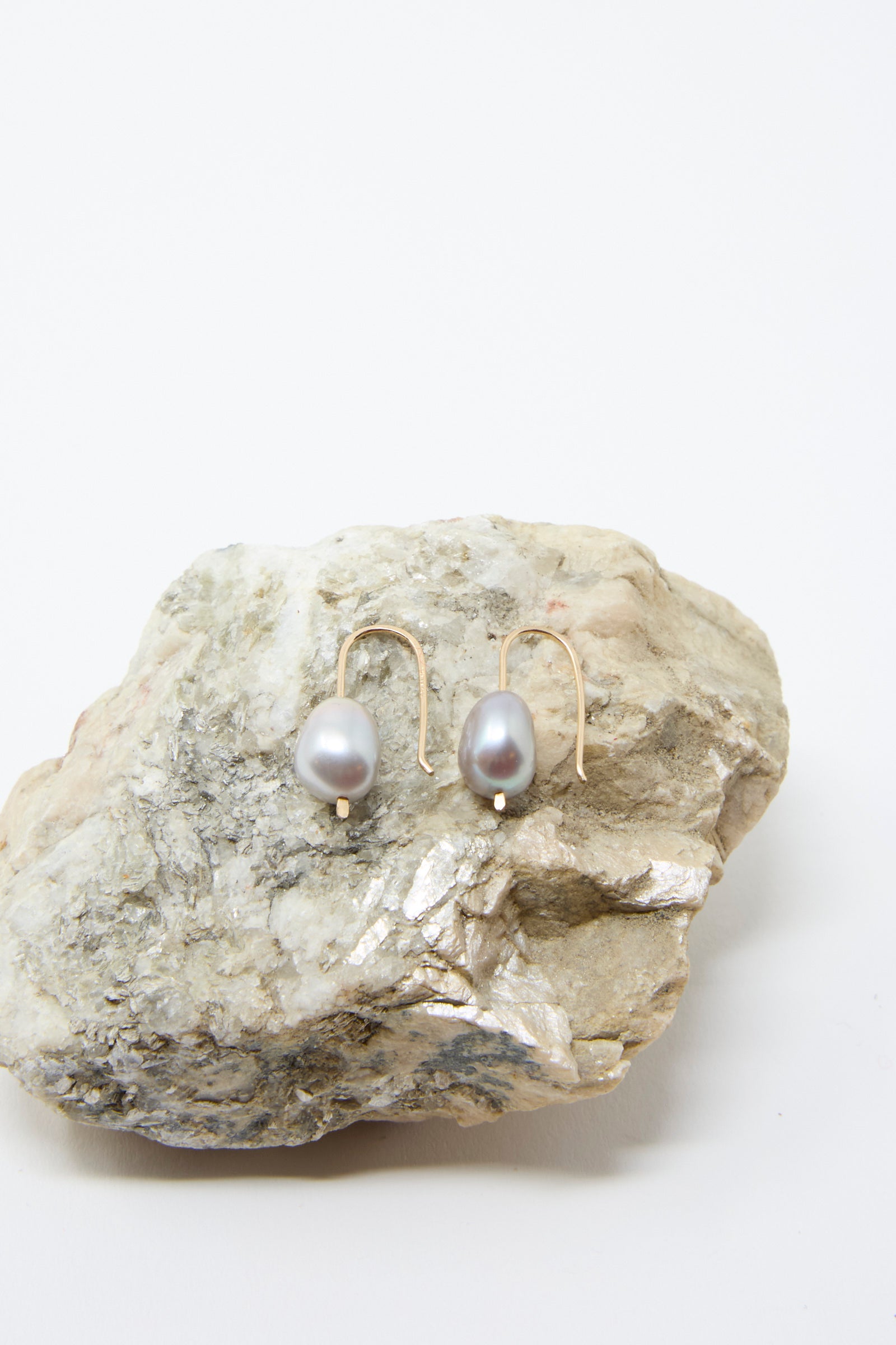 The Petite Baroque Pearl Drop Earrings in Silver by Mary MacGill, crafted with hammered gold-filled wire and adorned with Baroque pearls, rest on a rough, light-colored stone against a simple background.