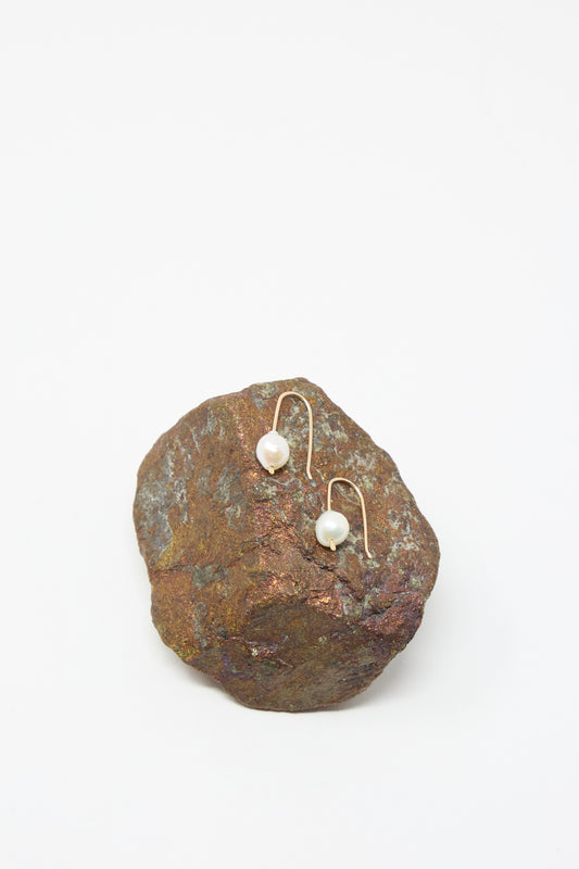 The Petite Baroque Pearl Drop Earrings in White by Mary MacGill, featuring curved gold-filled hooks, are displayed on a rough brown rock against a white background.