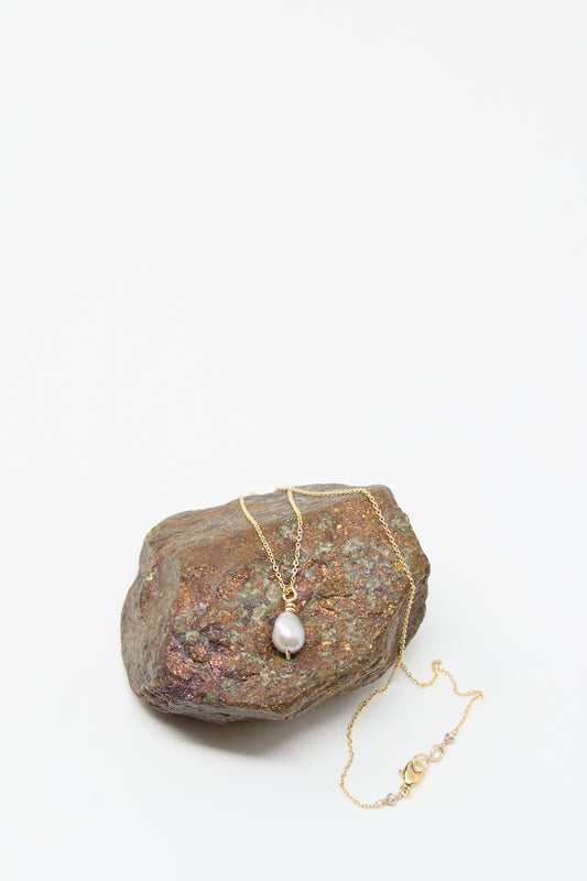 The Mary MacGill Petite Baroque Pearl Drop Necklace in Silver, featuring a single baroque pearl pendant, is draped over a brown stone against a plain white background.