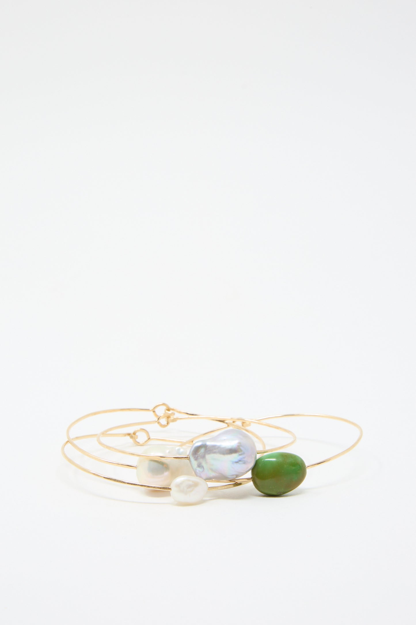 The Stone Cuff in Turquoise by Mary MacGill is featured with three gold hoop bracelets crafted with gold-filled wire, highlighting an iridescent bead, a white bead, and a captivating green bead evocative of a Turquoise stone, all elegantly displayed on a white background.