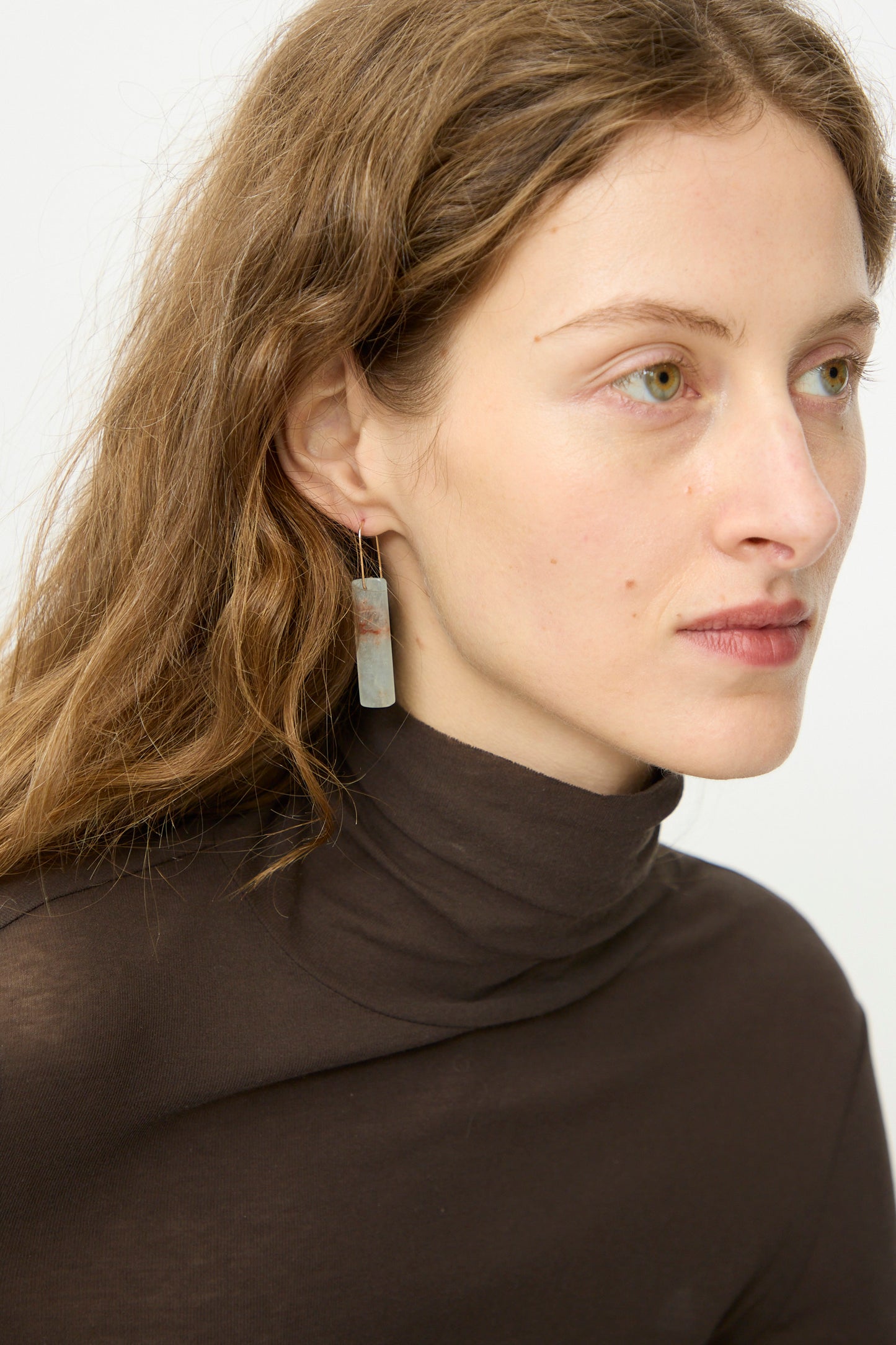 A woman with light skin and wavy brown hair, dressed in a brown turtleneck, wears Mary MacGill Stone Drop Earrings in Blue Calcite Rectangular Stone while glancing to the side.