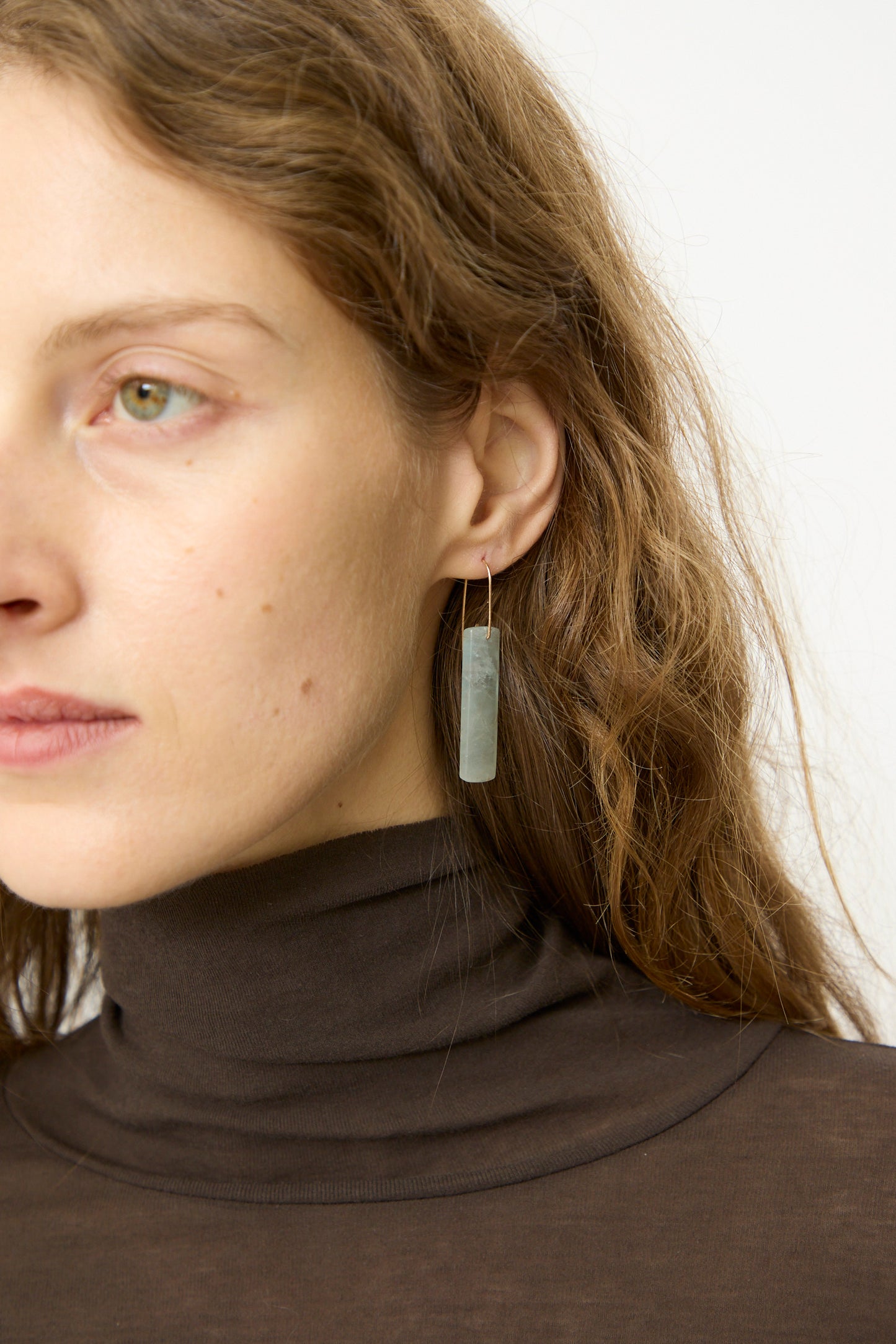 A woman with light skin and wavy brown hair wears a brown turtleneck and Mary MacGill's Stone Drop Earrings featuring rectangular Blue Calcite stones, shown from the shoulders up against a plain backdrop.