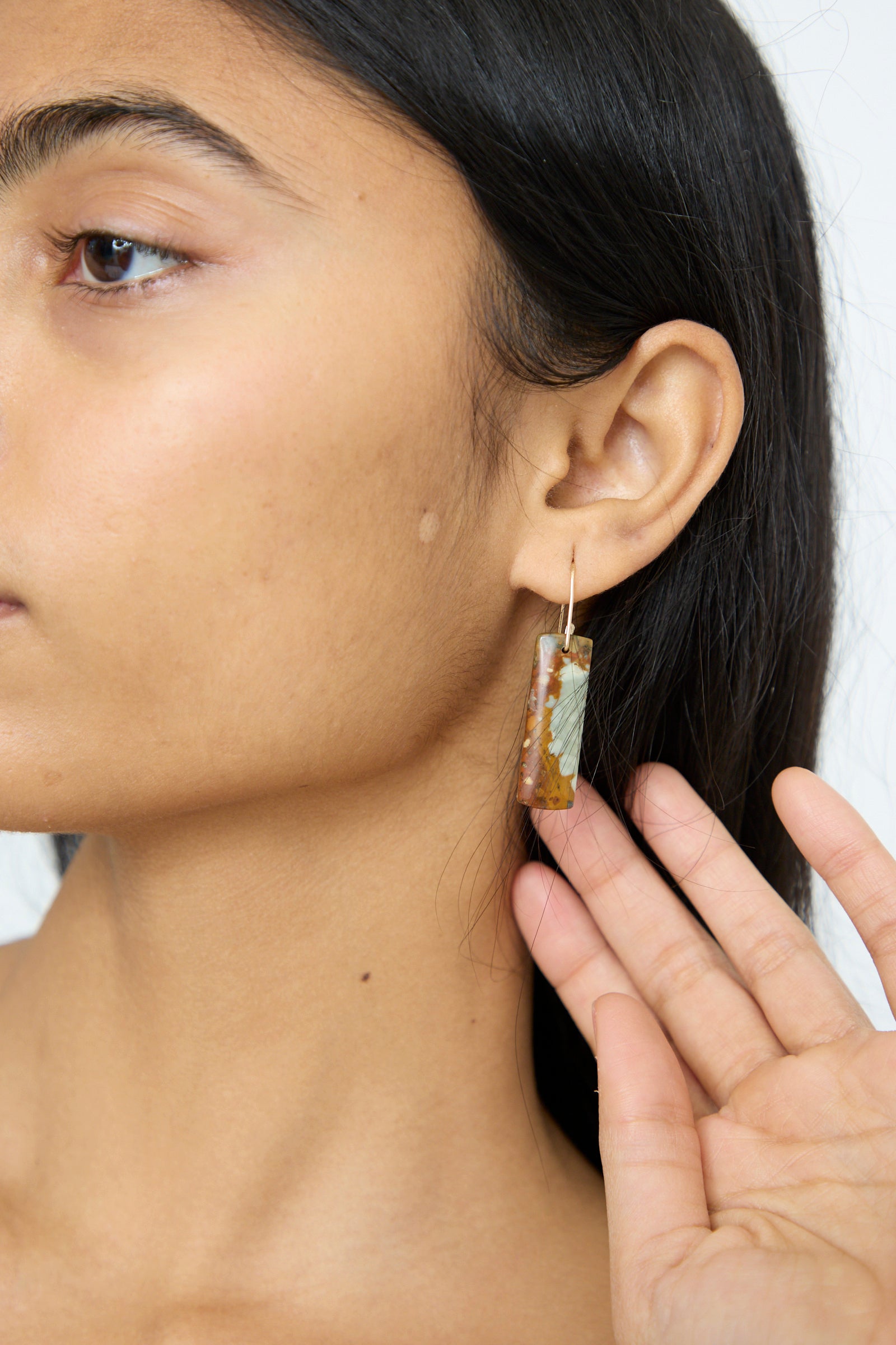 A person with long dark hair holds a rectangular Stone Drop Earring in Jasper, made by Mary MacGill with delicate handmade gold-filled wire.