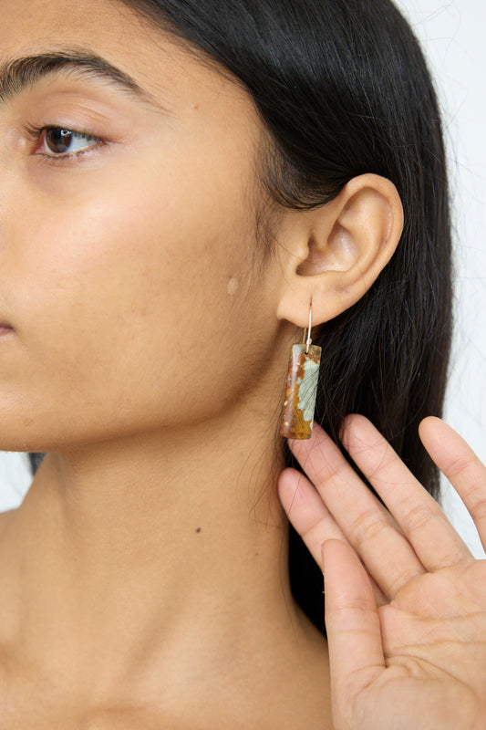 A person with long dark hair holds a rectangular Stone Drop Earring in Jasper, made by Mary MacGill with delicate handmade gold-filled wire.