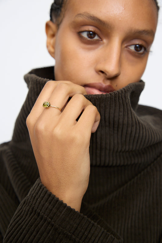 A person in a dark ribbed turtleneck holds up the collar, showcasing Mary MacGill’s 14K Floating Ring in Petite Green Tourmaline.