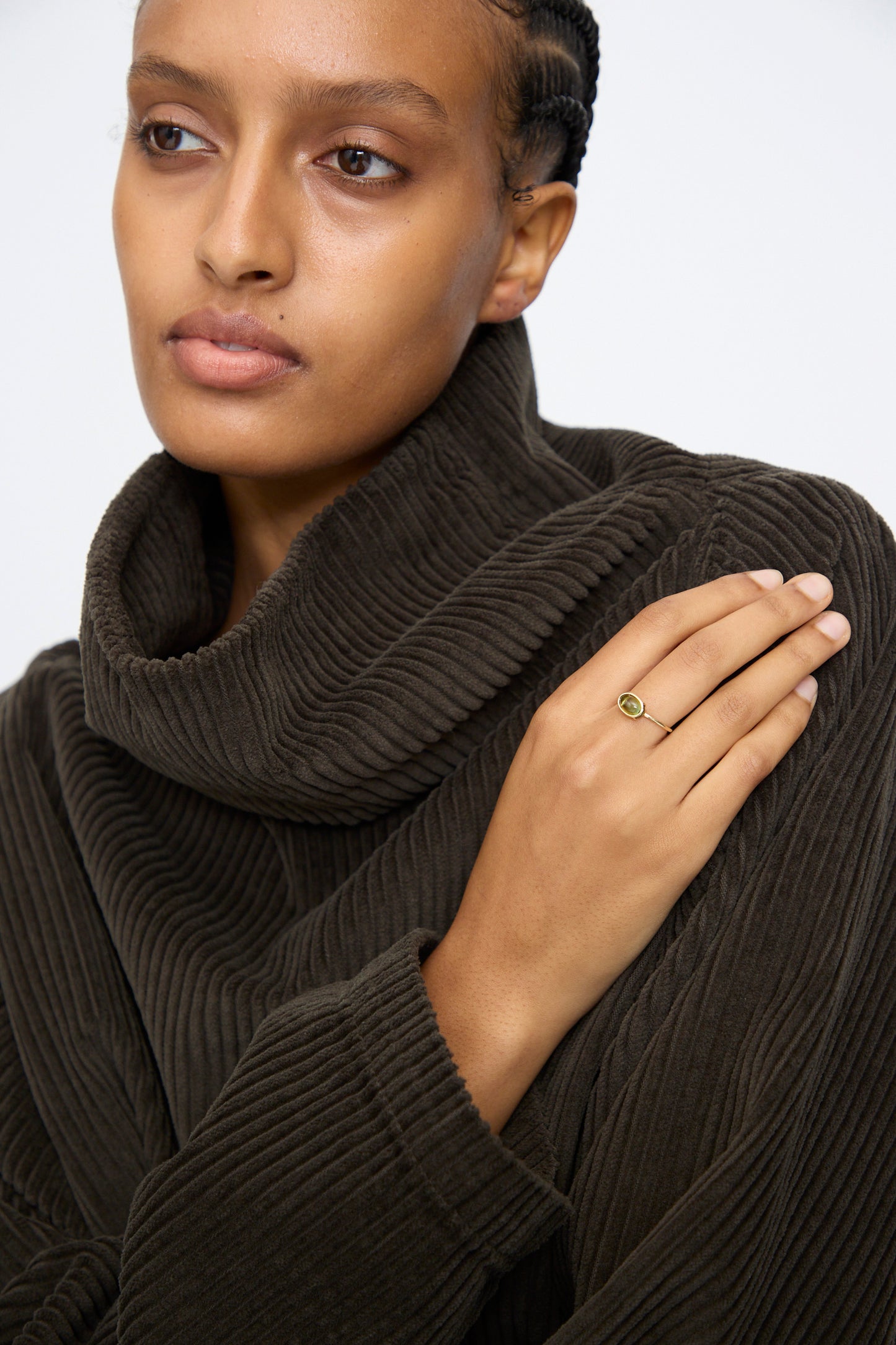 A person wearing a dark brown ribbed turtleneck rests their hand on their shoulder, showcasing the Mary MacGill 14K Floating Ring in Petite Green Tourmaline.