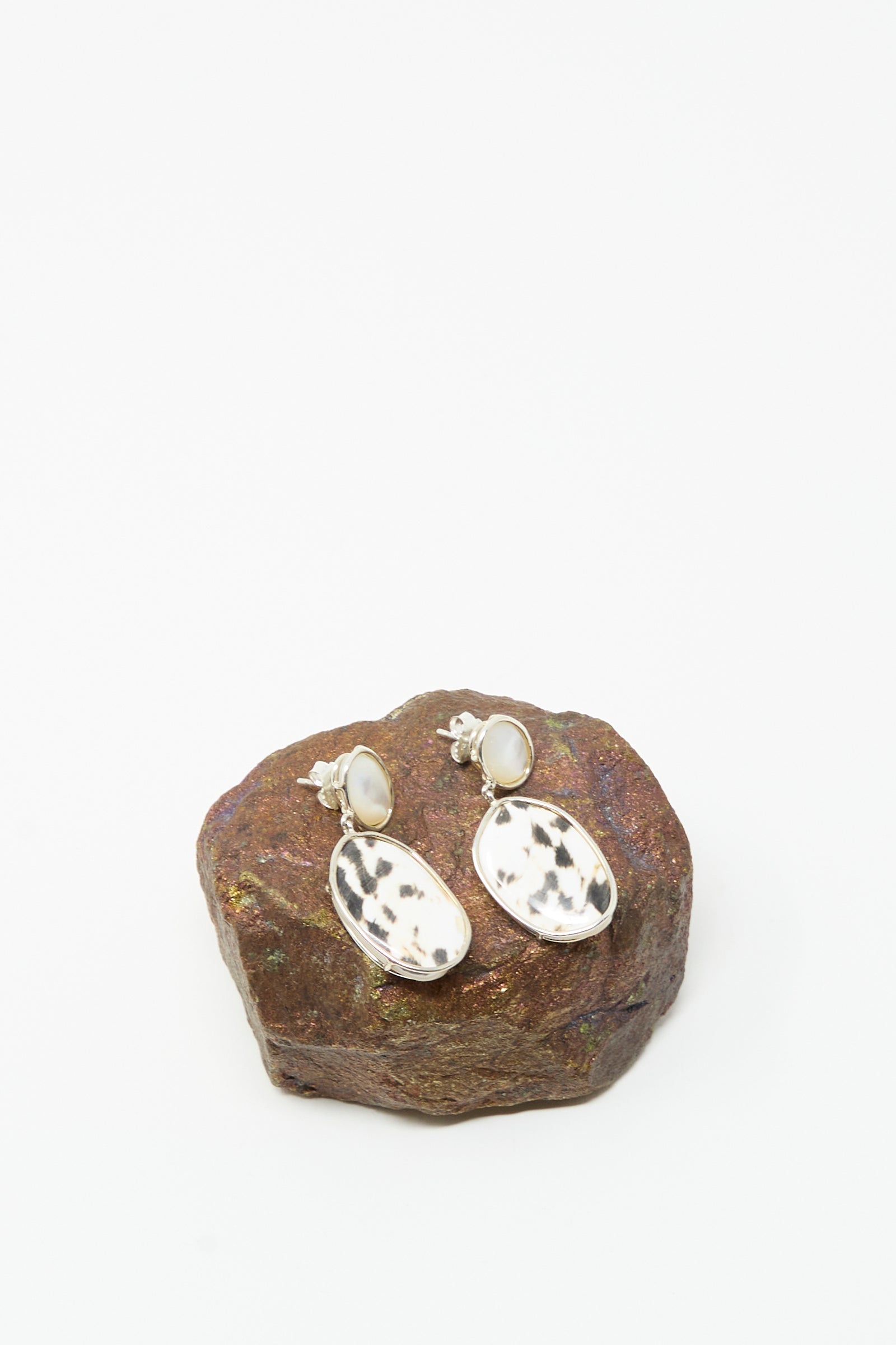Mary MacGill Double Stone Drop Earrings in Moonstone and Cowrie Shell, featuring oval cowrie shell pendants and white moonstone studs, are displayed on a brown rock against a white background.