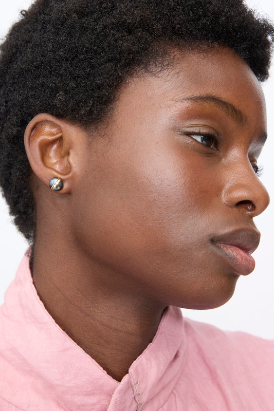 A person wearing the handmade Grande Baroque Pearl Studs in Charcoal, featuring a charcoal baroque pearl with a gold band.