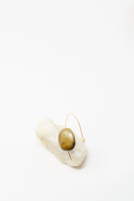 The Stone Cuff Bracelet in Brown Turquoise Stone by Mary MacGill, a gold-filled handmade piece, features an oval brown stone and is displayed on a white textured rock against a plain white background.