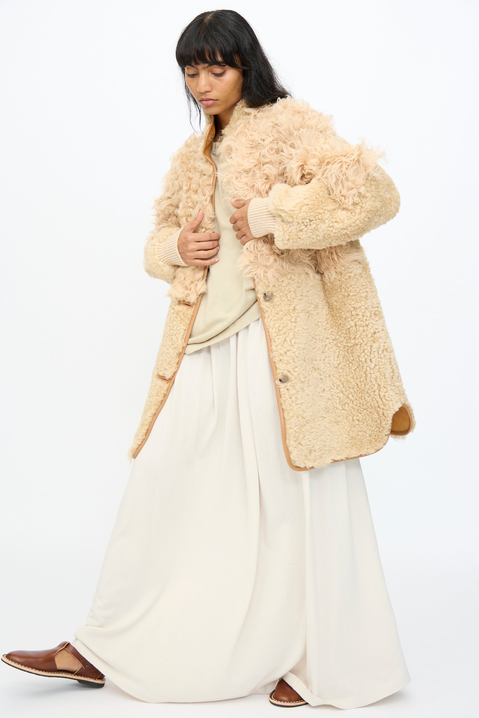 A person wears the Milena Silvano Allie Coat in Oat—a cream, textured patchwork sheepskin coat made in Turkey—paired with a light shirt, wide cream pants, and brown slip-on shoes, standing against a plain white background.