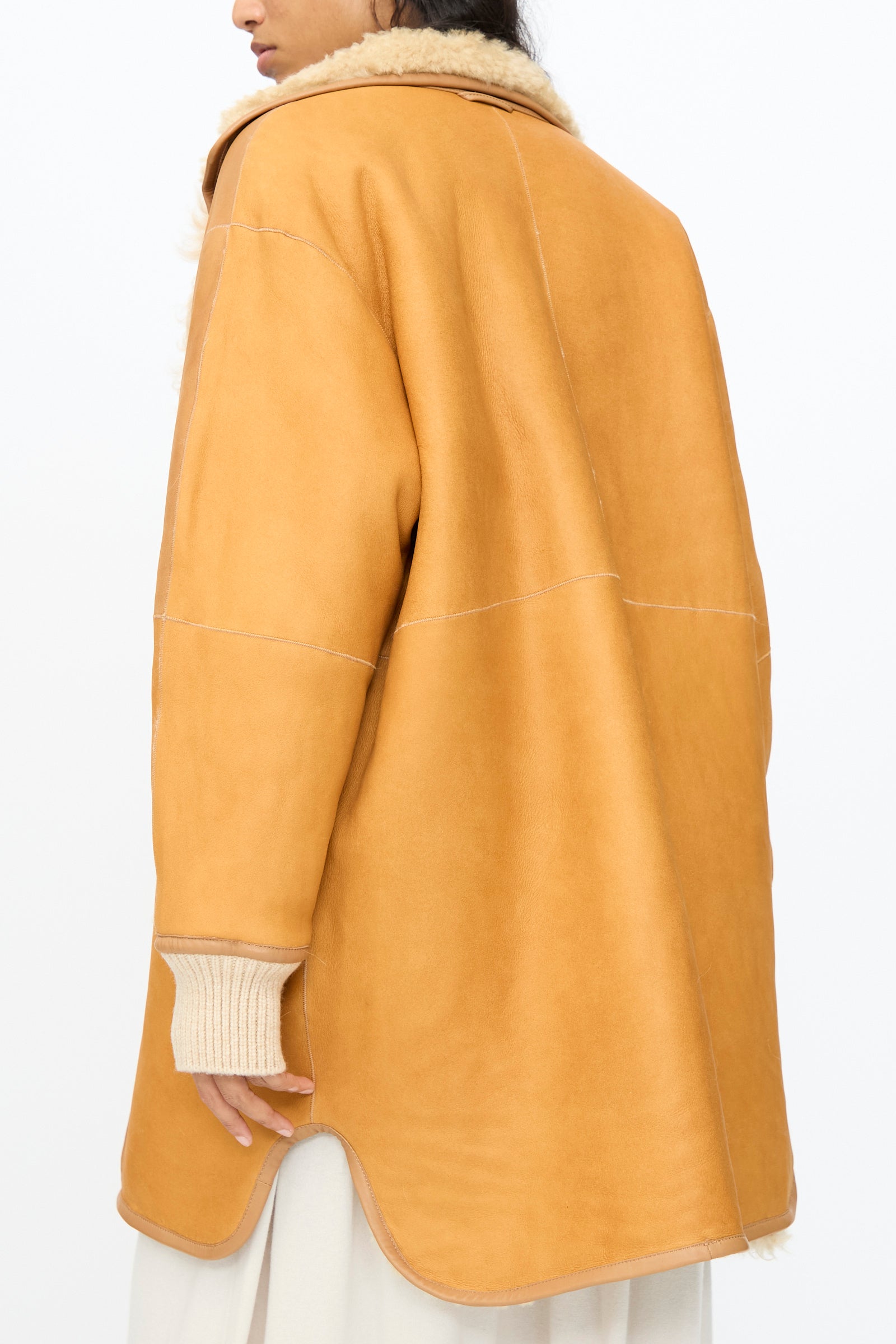 A person wears the Milena Silvano Allie Coat in Oat, featuring visible stitching and knit sleeves, photographed from behind against a plain white background.