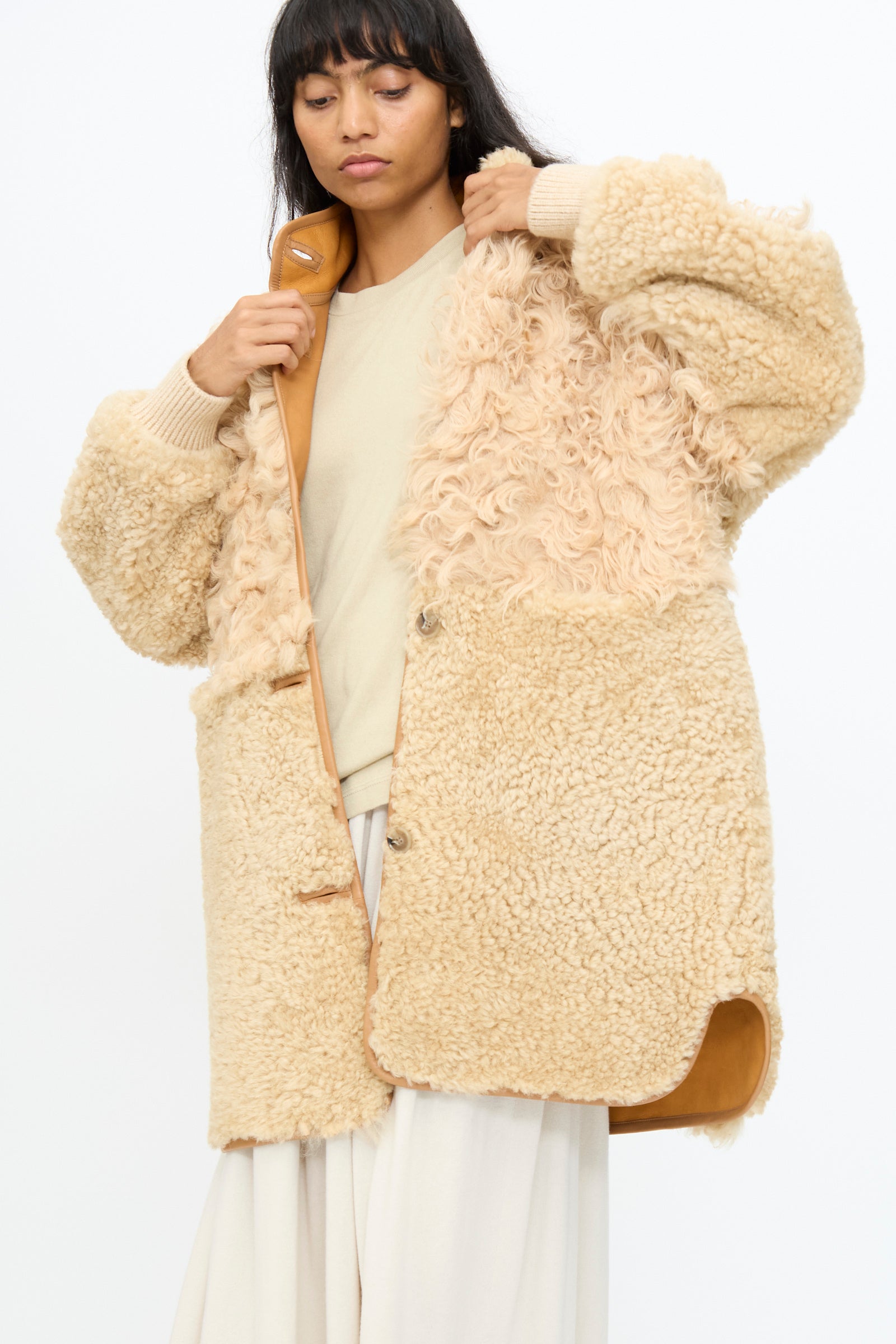 A woman wears the Milena Silvano Allie Coat in Oat—an oat-colored patchwork sheepskin coat made in Turkey—over a light sweater and wide-leg pants, standing against a plain white background.