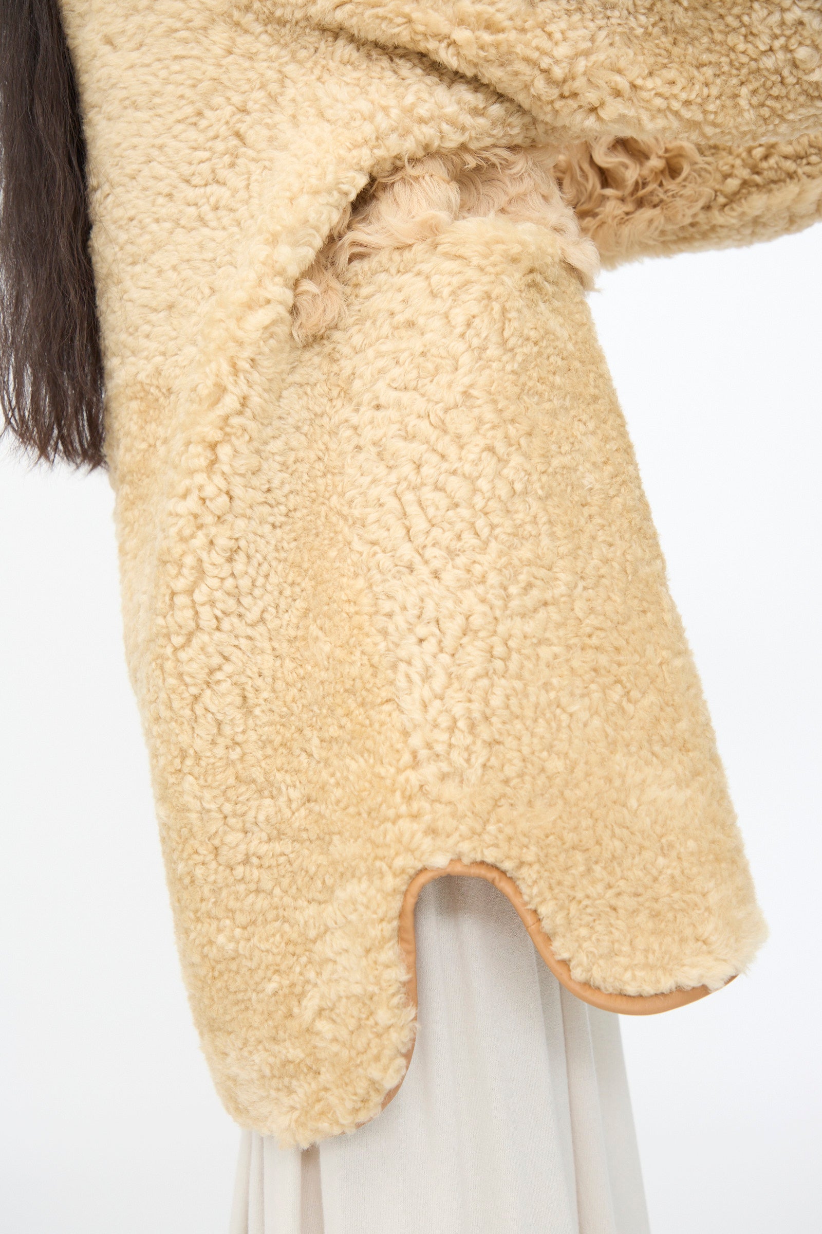 Close-up of a person wearing the Milena Silvano Allie Coat in Oat, a beige, textured patchwork sheepskin piece, over light skirt or pants; long dark hair visible on the left.