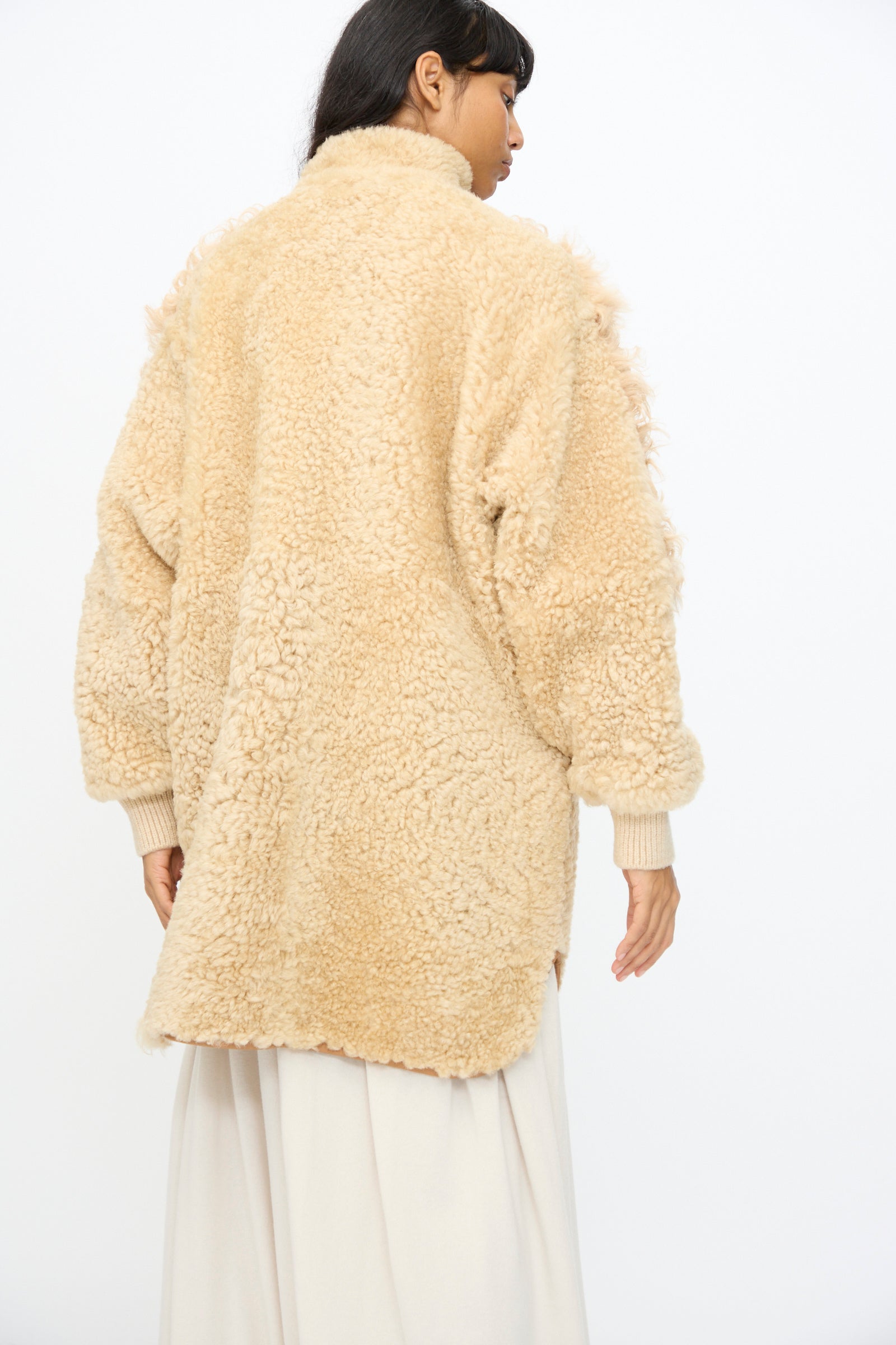 A person is seen from behind wearing the Milena Silvano Allie Coat in Oat—an oversized, textured patchwork sheepskin coat—over a long, light skirt, set against a plain background.