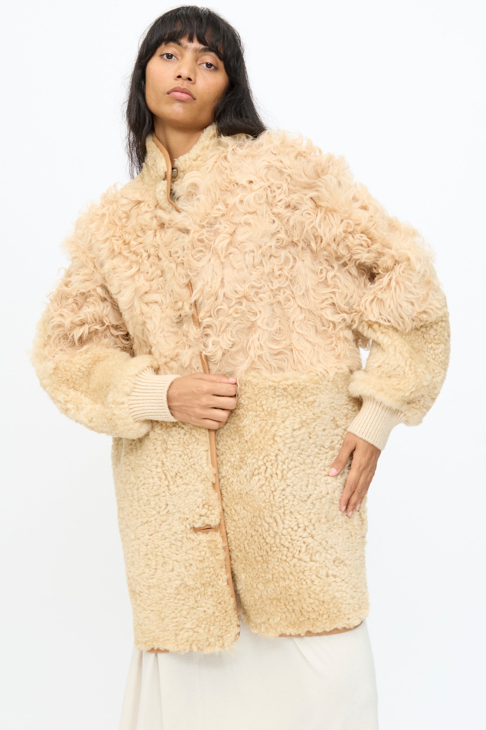 A woman wears the Milena Silvano Allie Coat in Oat, a light beige patchwork sheepskin piece with curly and smooth textures, made in Turkey, standing against a plain white background.