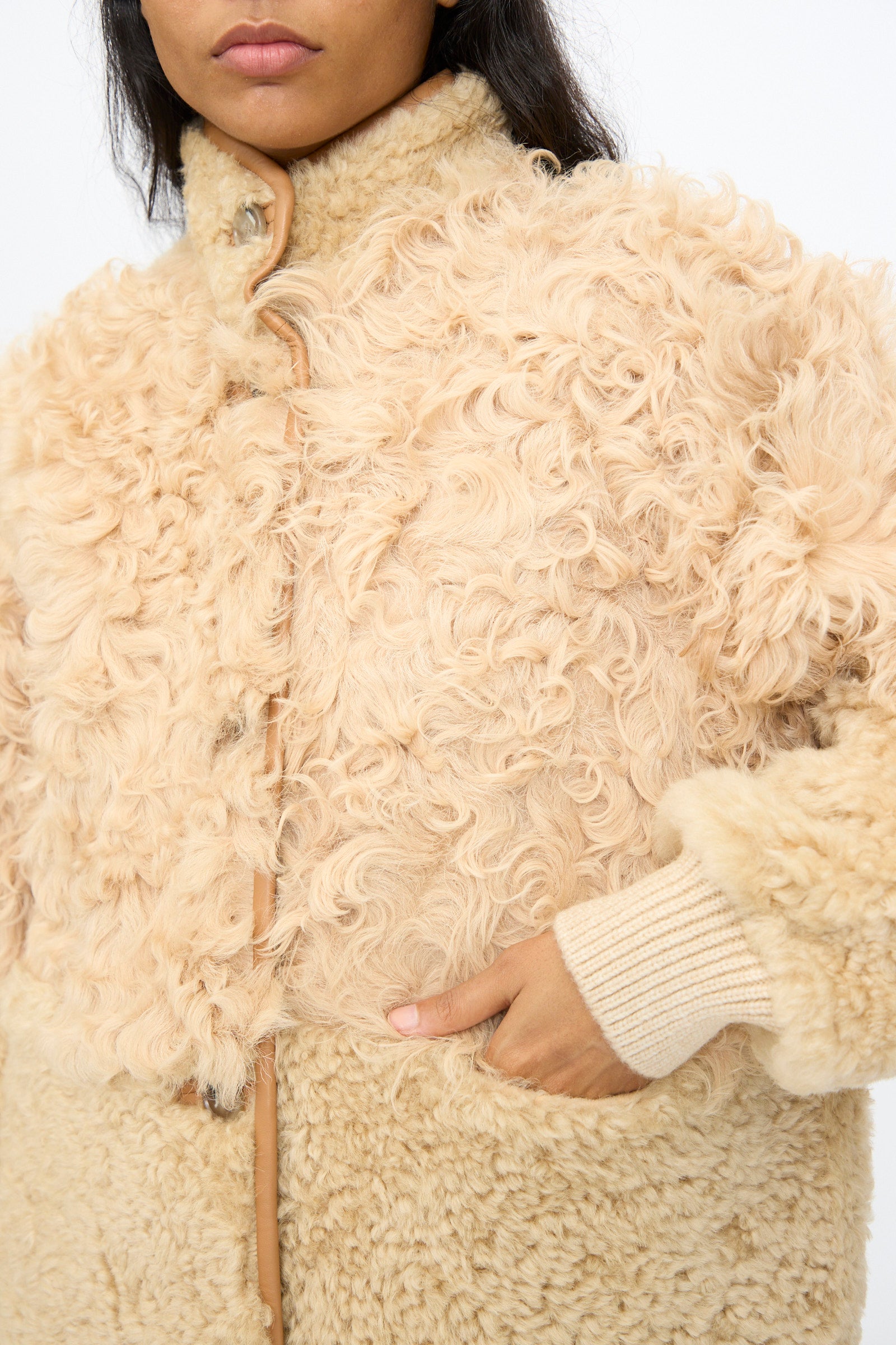 A person wearing the Milena Silvano Allie Coat in Oat—a beige, curly-textured patchwork sheepskin coat made in Turkey—with one hand in the pocket.