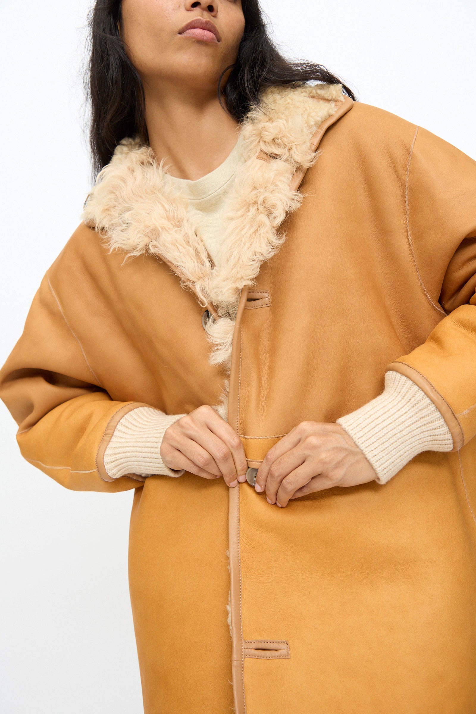 A person fastens the Milena Silvano Allie Coat in Oat—a tan patchwork sheepskin style with cream cuffs—against a plain white background.
