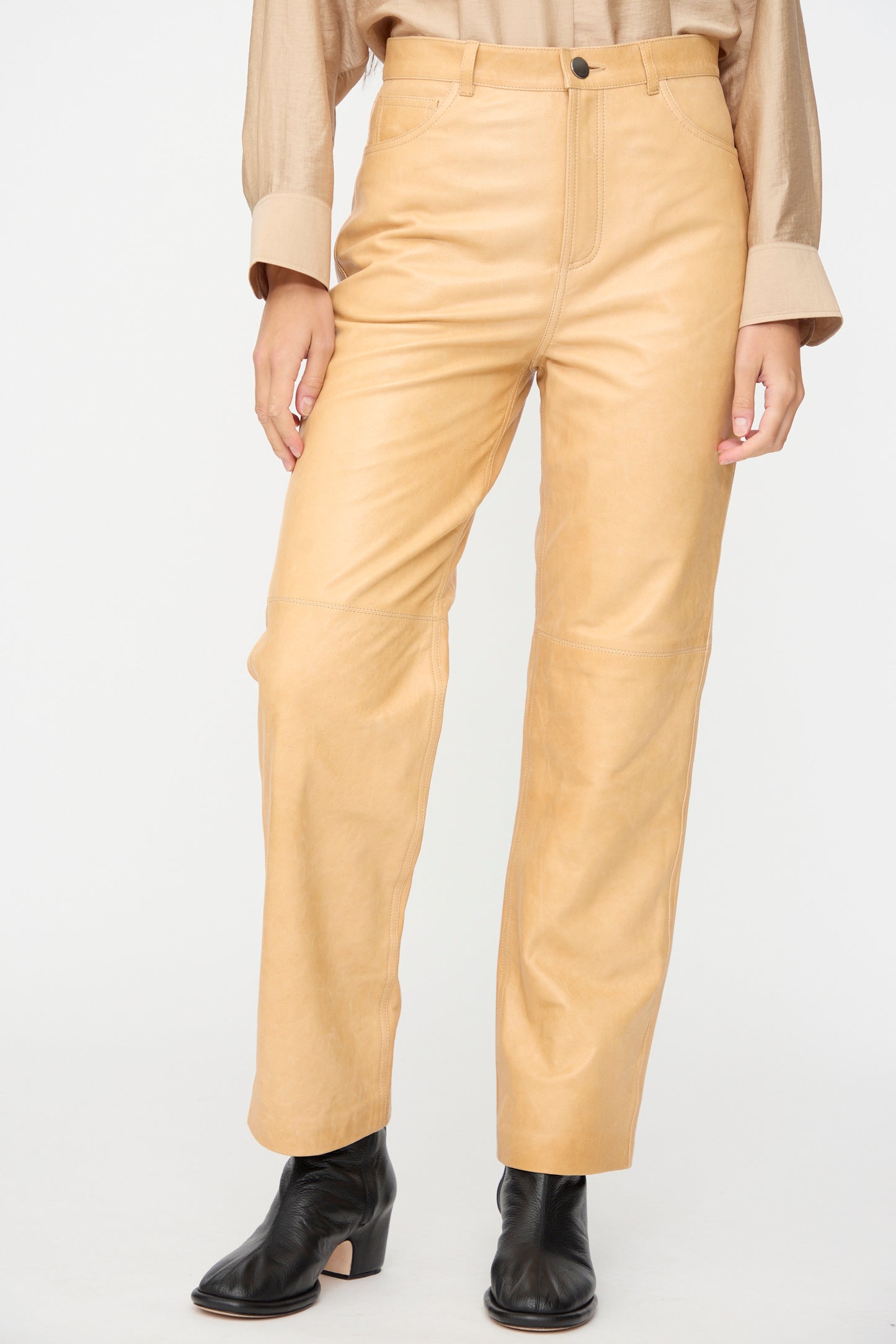 A person wearing Milena Silvano’s Leather 5 Pocket Trousers in Bare, paired with a beige button-up shirt and black ankle boots, standing against a plain white background.