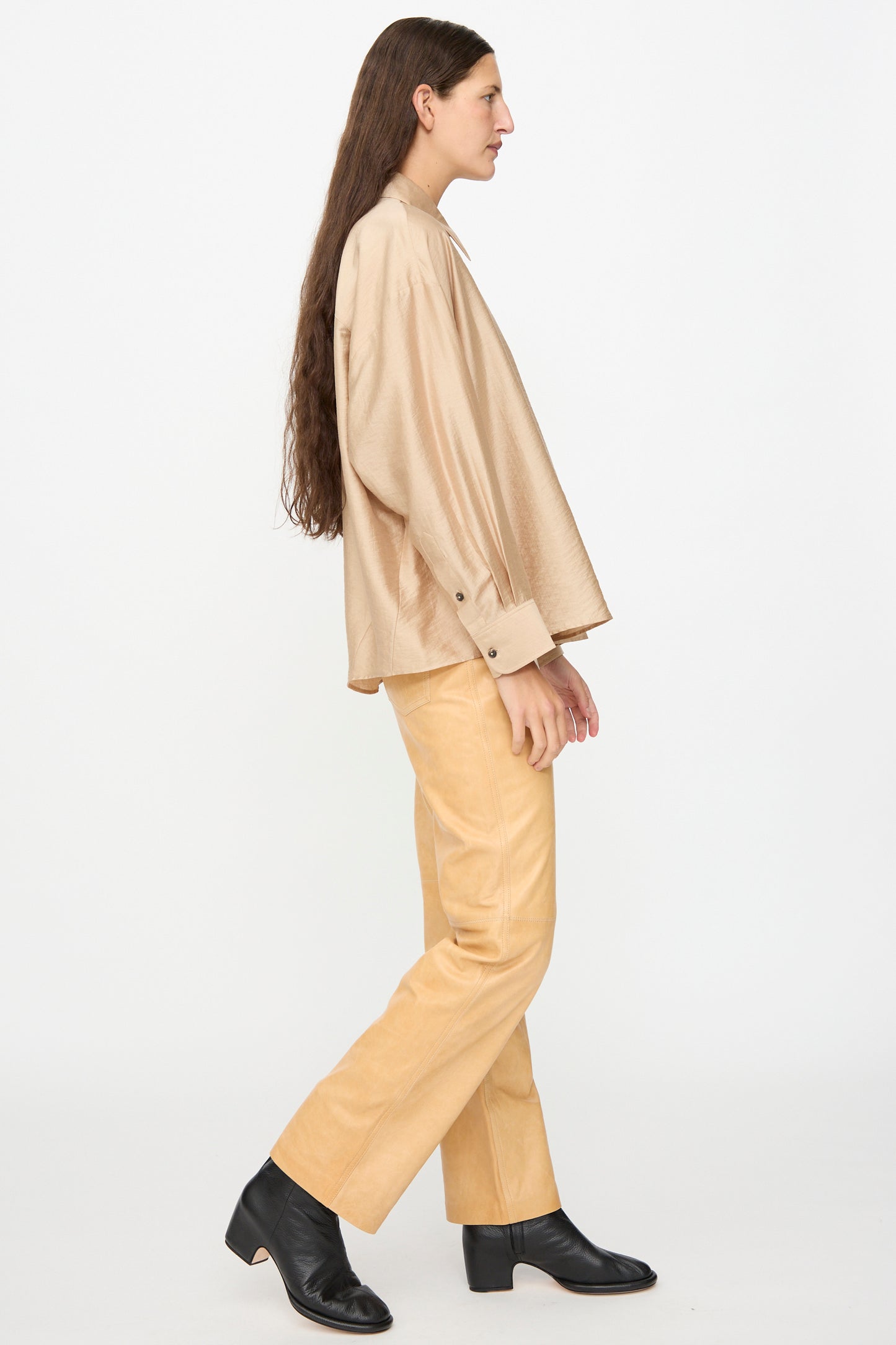 A person with long brown hair stands sideways against a plain white background, wearing Milena Silvano’s Leather 5 Pocket Trousers in Bare, paired with a loose beige shirt and black vegetable tanned leather shoes.