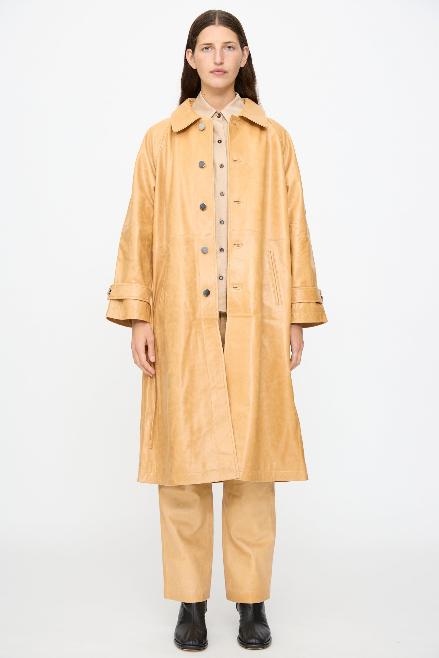 A person stands against a white background, wearing the Milena Silvano Leather Maria Trench in Bare over matching pants and shirt, styled with black ankle boots.