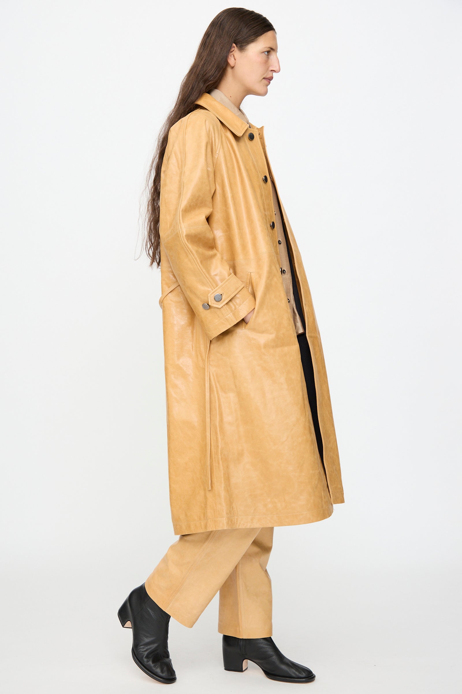 A person with long brown hair wears the Milena Silvano Leather Maria Trench in Bare, matching pants, and black heeled boots while walking against a plain white background.