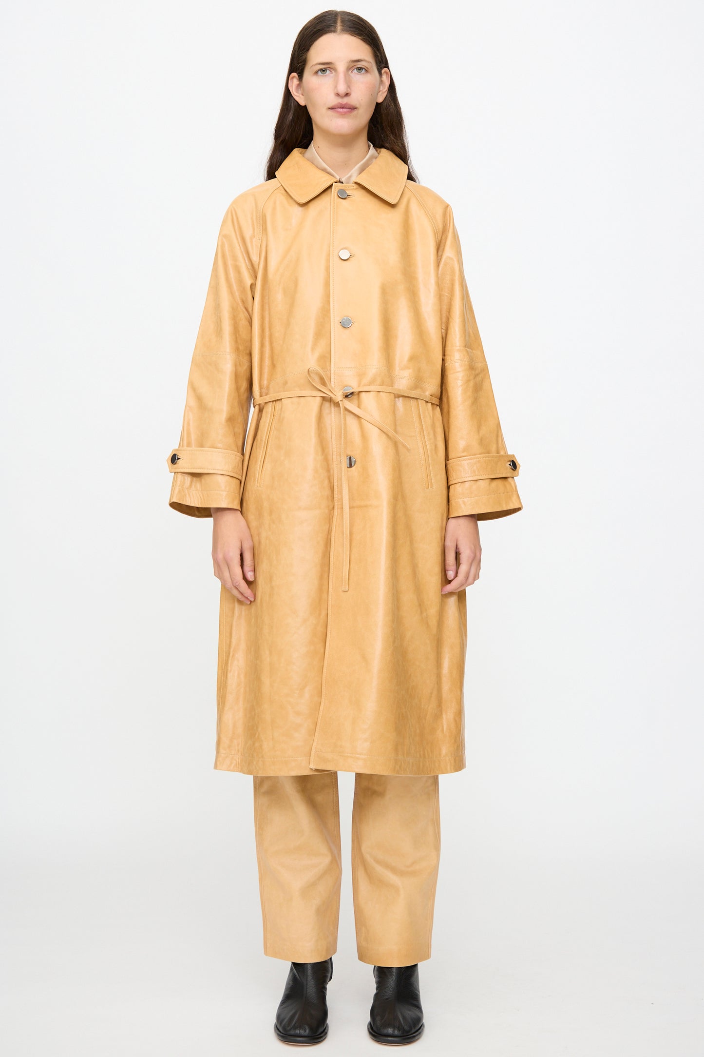 A person stands facing forward against a plain white background wearing the Milena Silvano Leather Maria Trench in Bare—a tan belted trench coat with a subtle shiny finish—paired with matching pants and black ankle boots.
