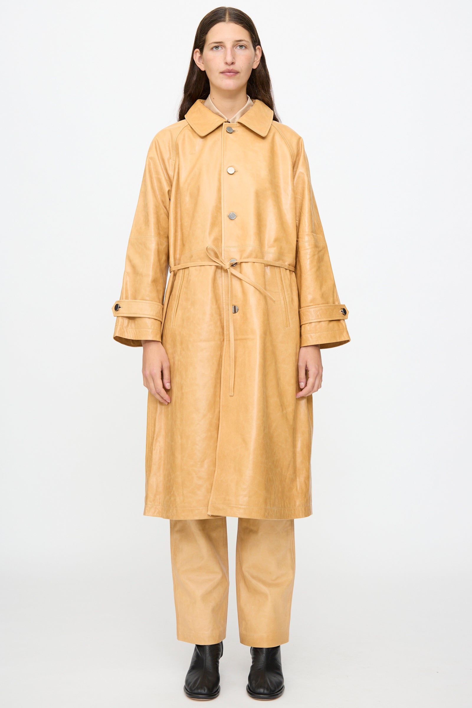 A person stands facing forward against a plain white background wearing the Milena Silvano Leather Maria Trench in Bare—a tan belted trench coat with a subtle shiny finish—paired with matching pants and black ankle boots.