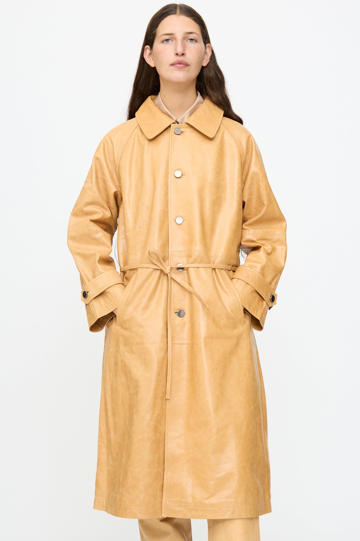 A model wears the Milena Silvano Leather Maria Trench in Bare—a tan, long-sleeve, button-up vegetable-tanned leather trench coat featuring a tie waist and subtle shiny finish—standing against a plain white background.