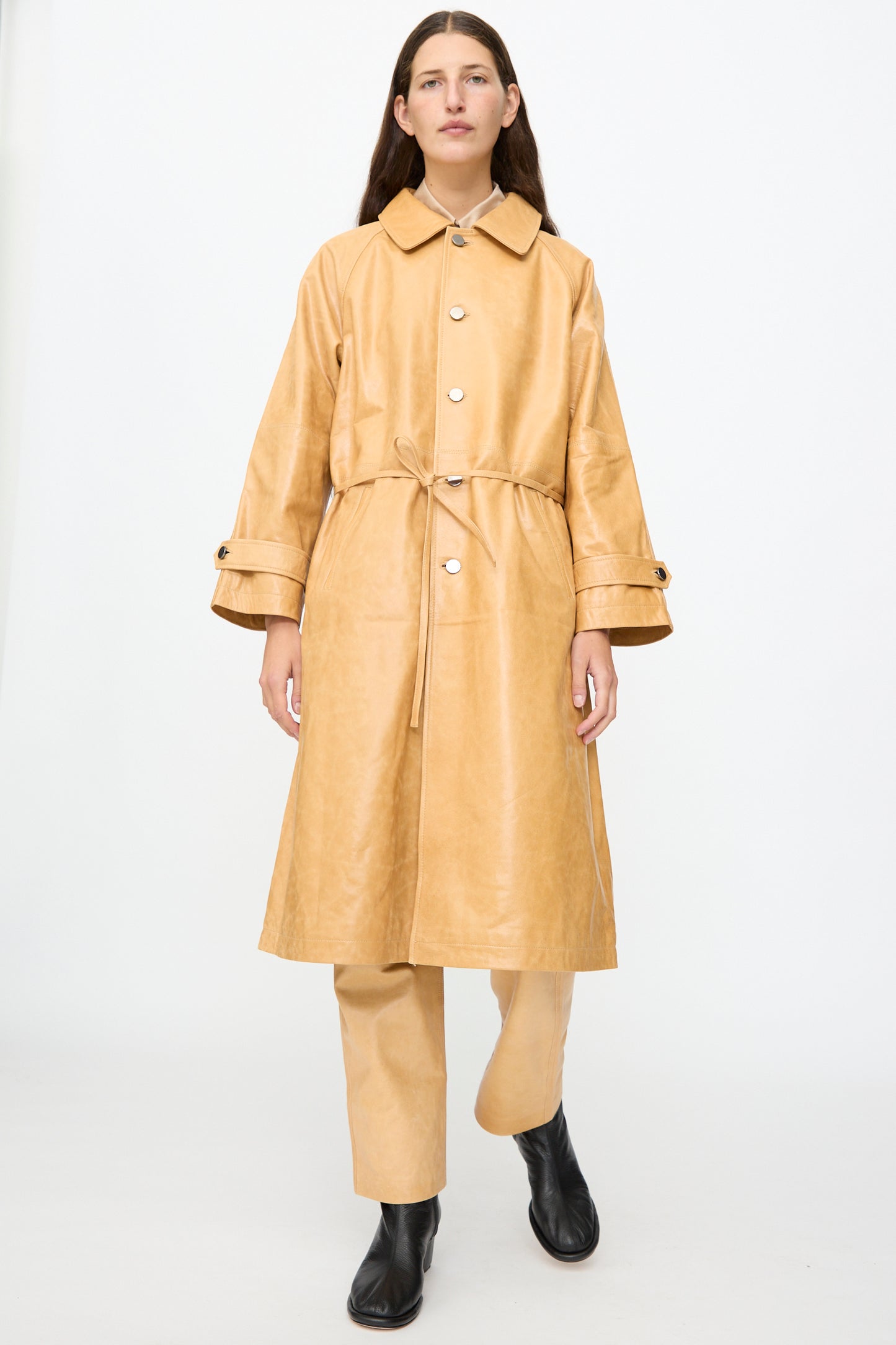Wearing the Milena Silvano Leather Maria Trench in Bare—a tan, waist-tied coat crafted from vegetable tanned leather with a subtle shiny finish—paired with matching tan pants and black boots, a person stands against a plain white background.