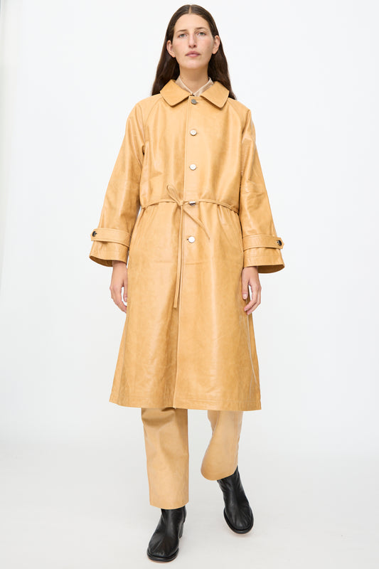 Wearing the Milena Silvano Leather Maria Trench in Bare—a tan, waist-tied coat crafted from vegetable tanned leather with a subtle shiny finish—paired with matching tan pants and black boots, a person stands against a plain white background.