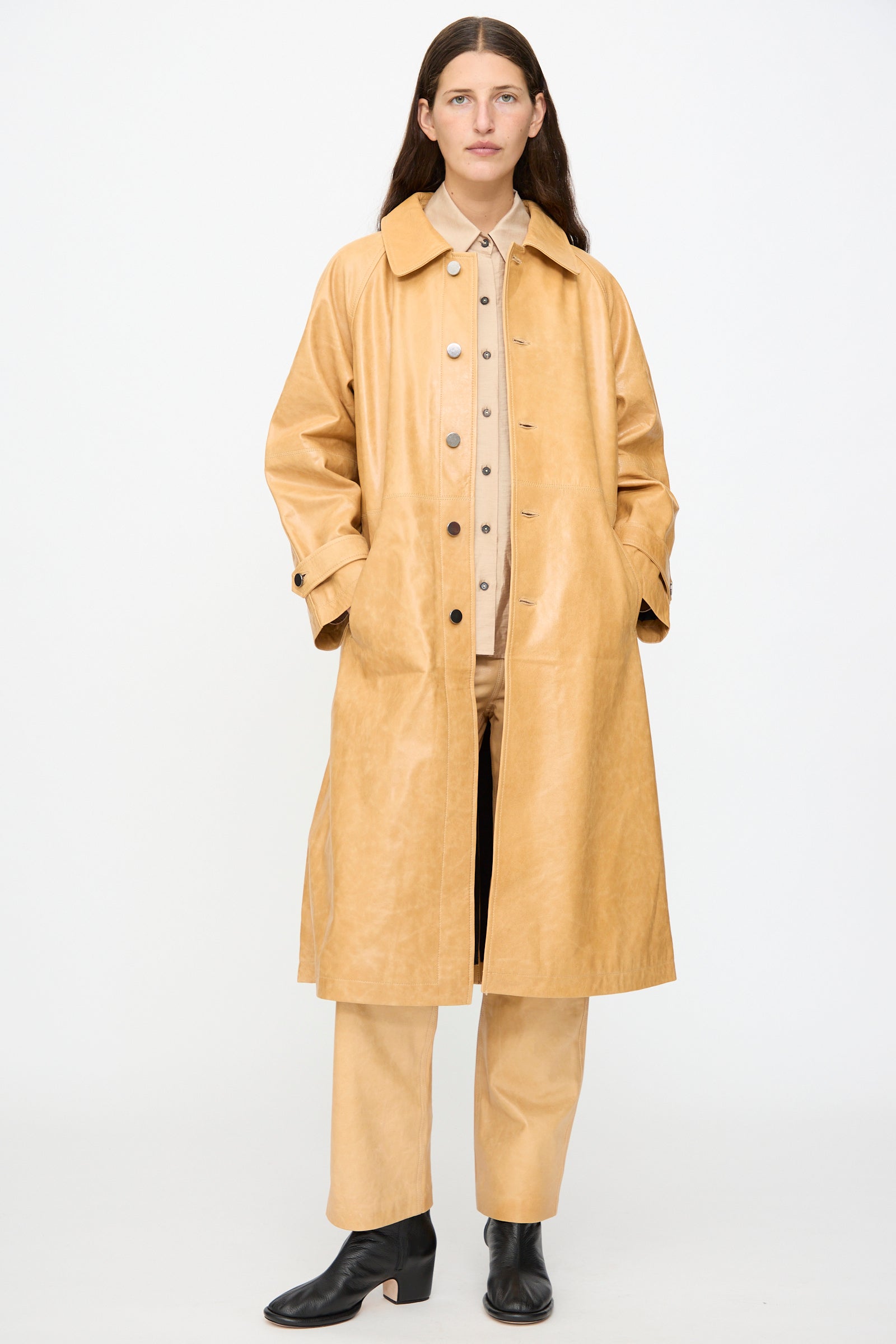 A person stands against a white background wearing the Leather Maria Trench in Bare by Milena Silvano, crafted from vegetable-tanned leather with a subtle shiny finish, paired with matching tan pants, a beige button-up shirt, and black ankle boots.