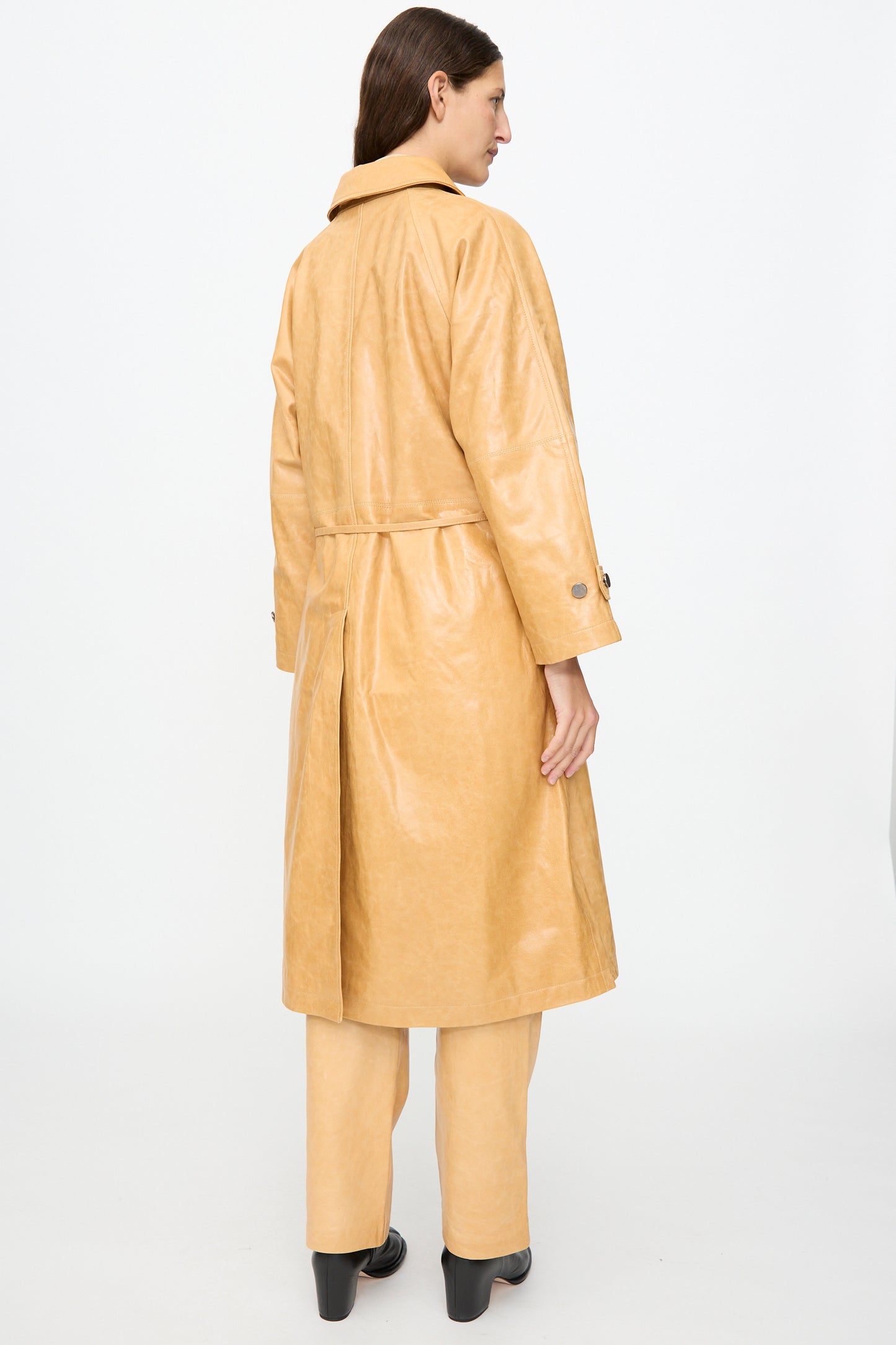 A person stands with their back to the camera on a plain white background, wearing the Milena Silvano Leather Maria Trench in Bare—crafted from vegetable-tanned leather with a subtle shiny finish—paired with matching pants and black boots.