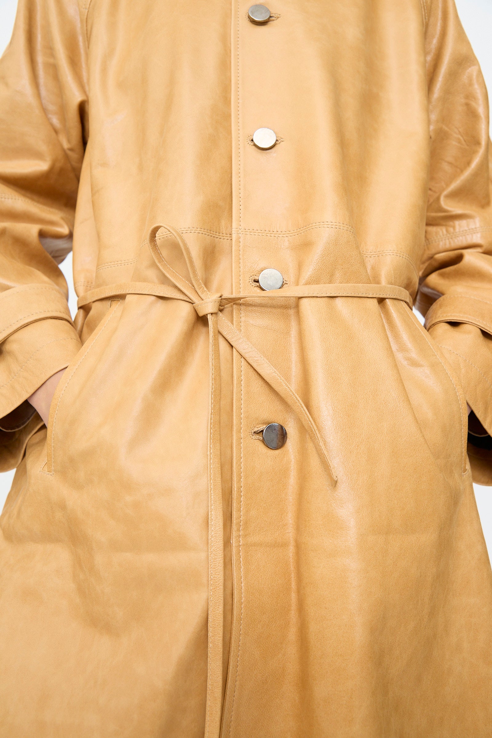 Close-up of the Milena Silvano Leather Maria Trench in Bare, crafted from tan vegetable-tanned leather with metallic buttons, a tied waist belt, and the wearer’s hands tucked into the pockets.