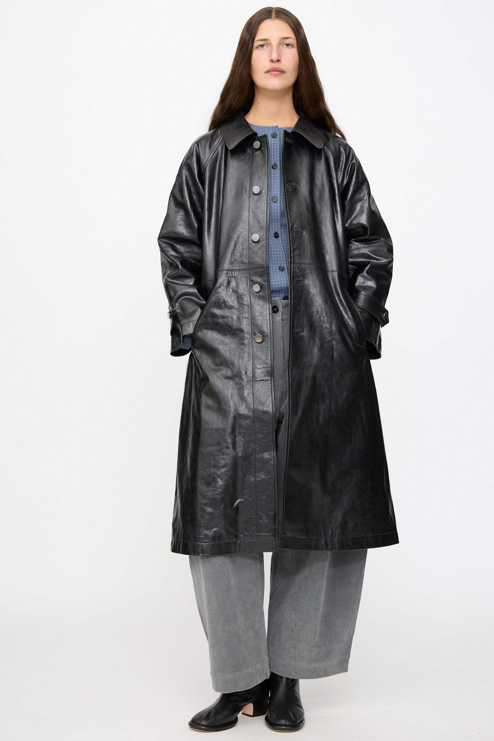 A person stands against a plain background, wearing the Milena Silvano Leather Maria Trench in Black, paired with a blue shirt, gray wide-leg pants, and black shoes.